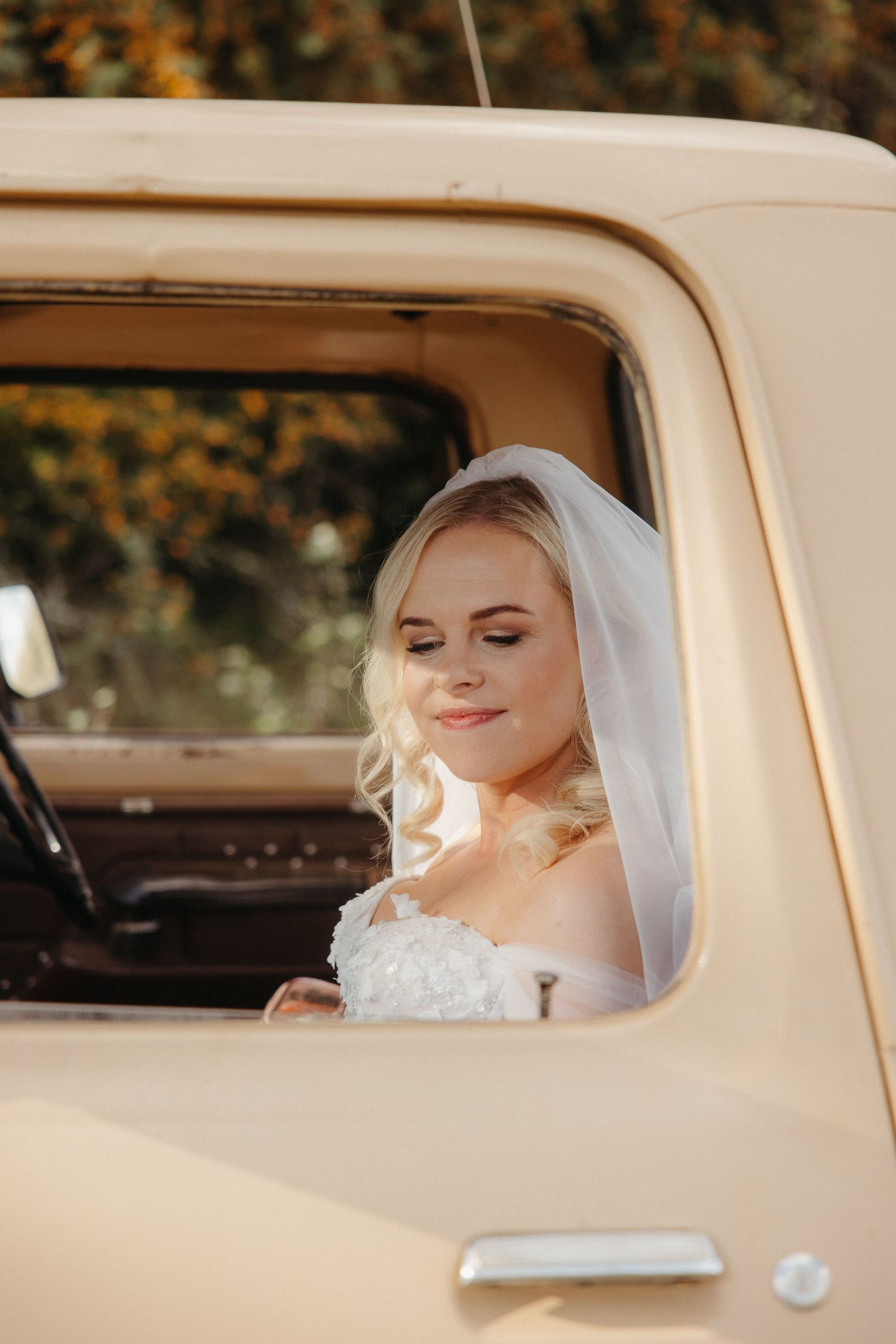 Bride in white gown and veil sitting inside a vintage cream truck, smiling softly on her wedding day.