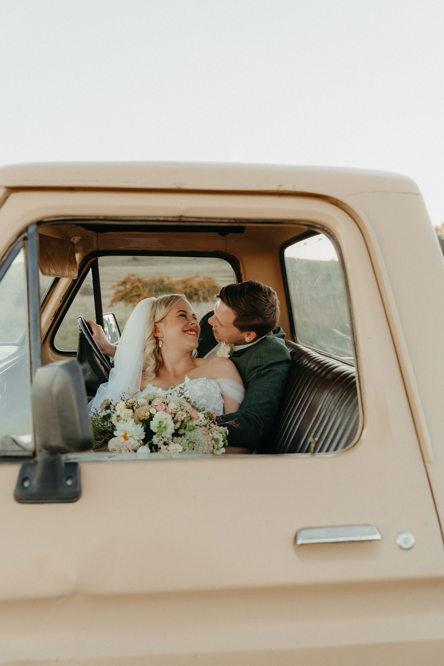 Bride and groom sharing a romantic moment inside a vintage beige truck, bride holding a pastel bouquet on their wedding day.