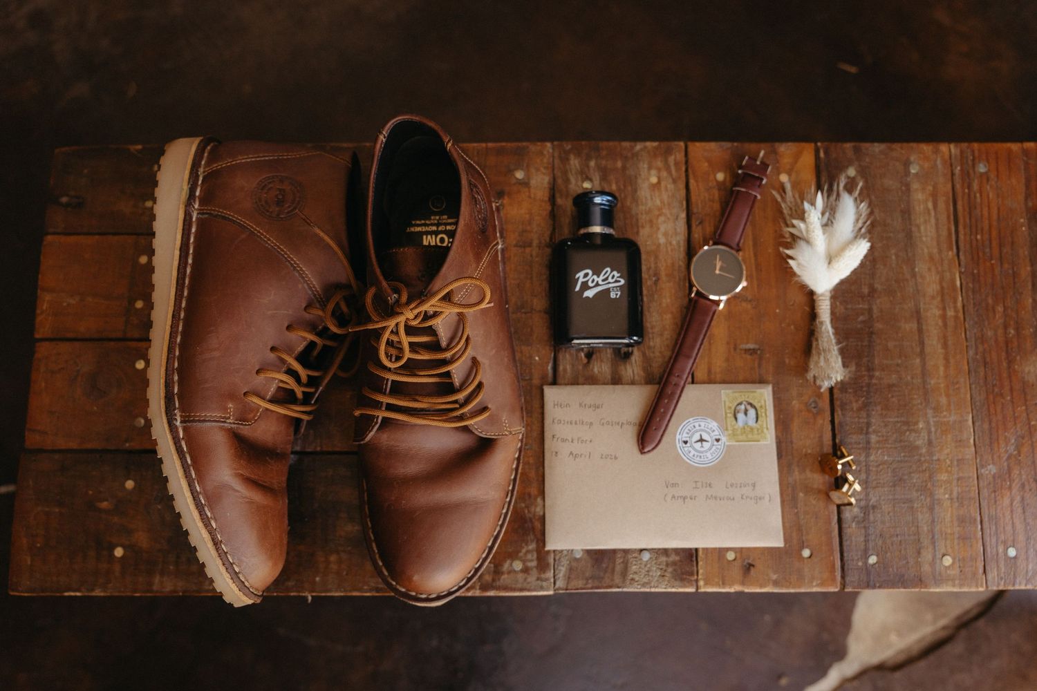 Brown leather lace-up dress shoes beside cologne, a watch, envelope, and feather on a wooden surface.