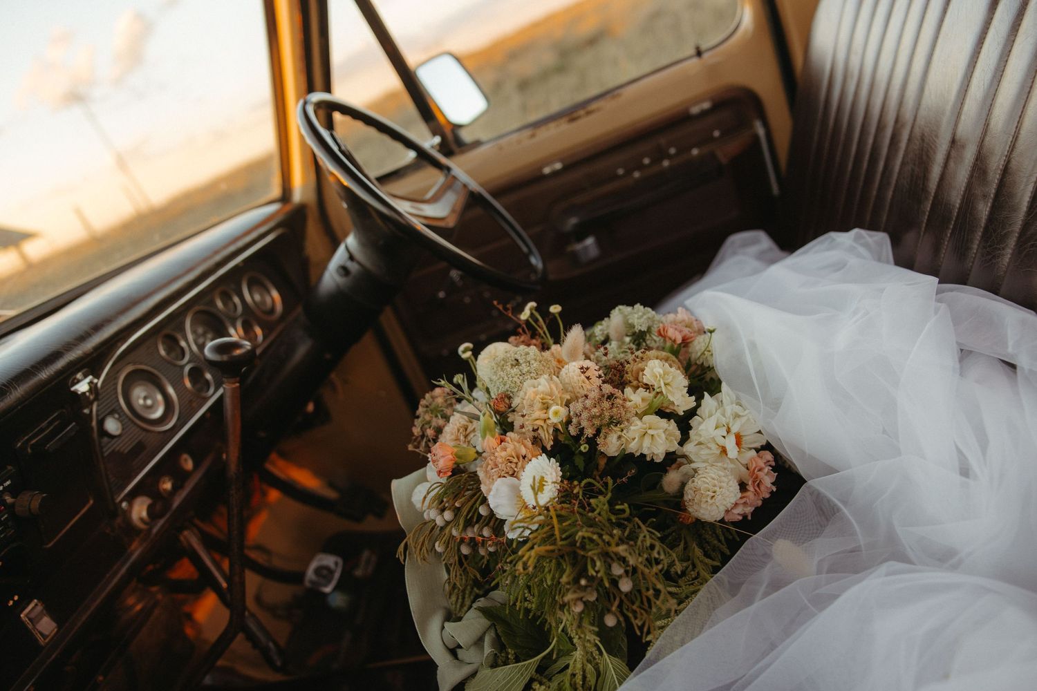 Bridal bouquet with pastel flowers resting on vintage truck seat beside white wedding dress at golden hour.