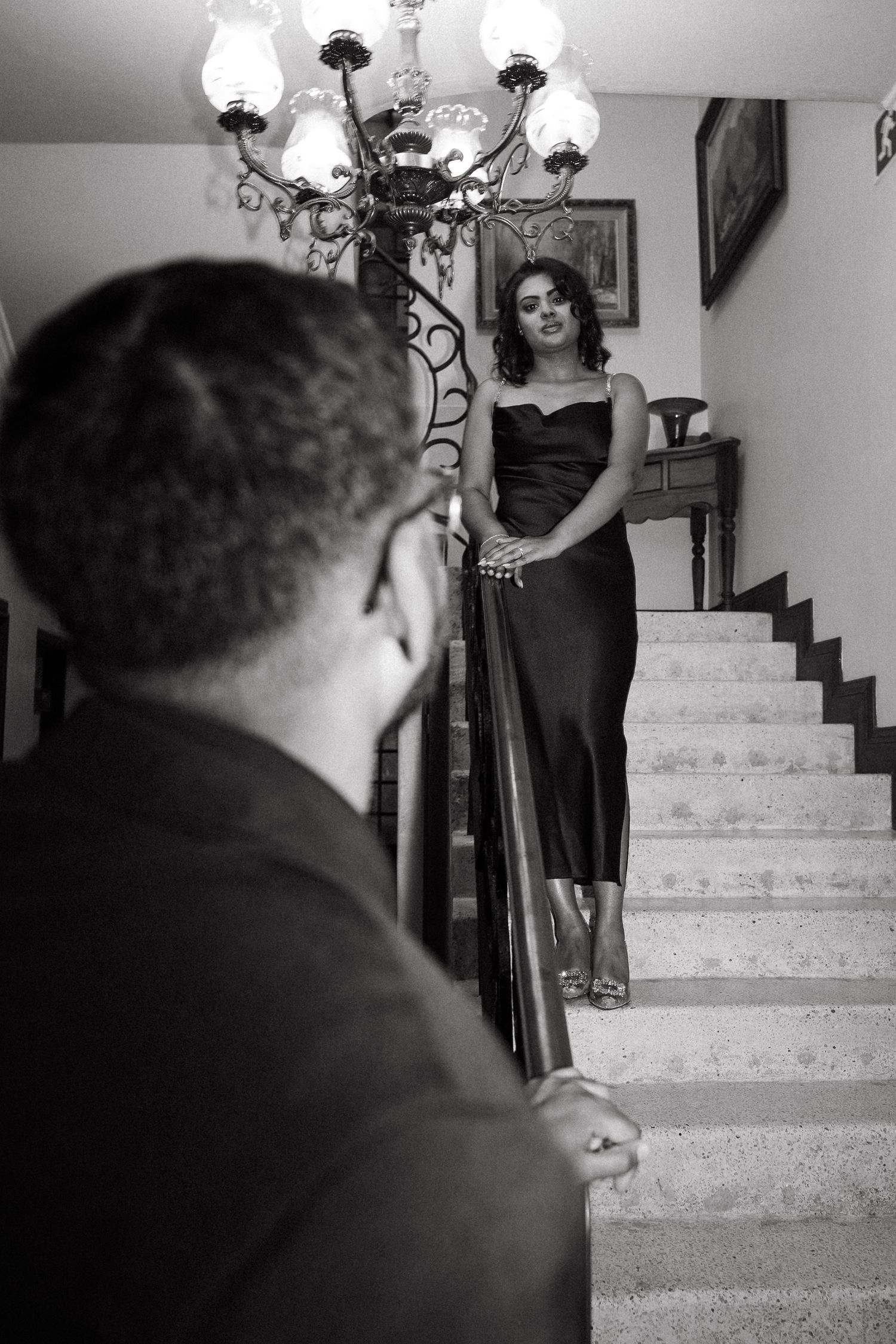 Black and white photo of elegant woman in satin dress descending staircase while man watches in formal attire.