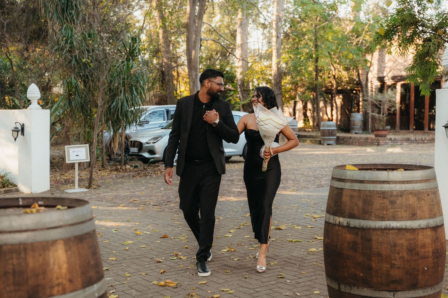 Couple walking together between wine barrels at an outdoor venue surrounded by trees.