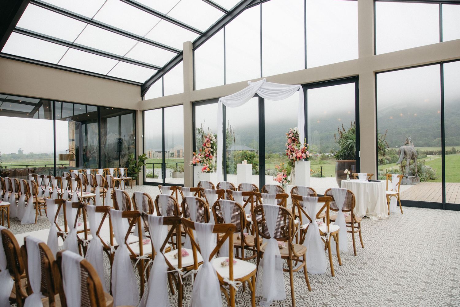 Elegant wedding ceremony with white draped chairs, floral arrangements, and glass walls overlooking misty mountains.