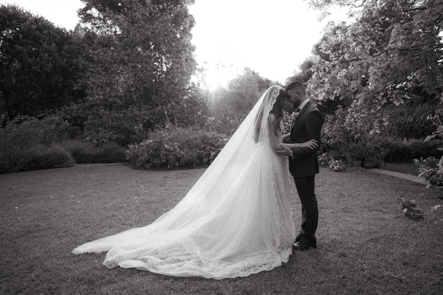 Black and white photo of bride and groom kissing in garden, long flowing veil trailing on grass.