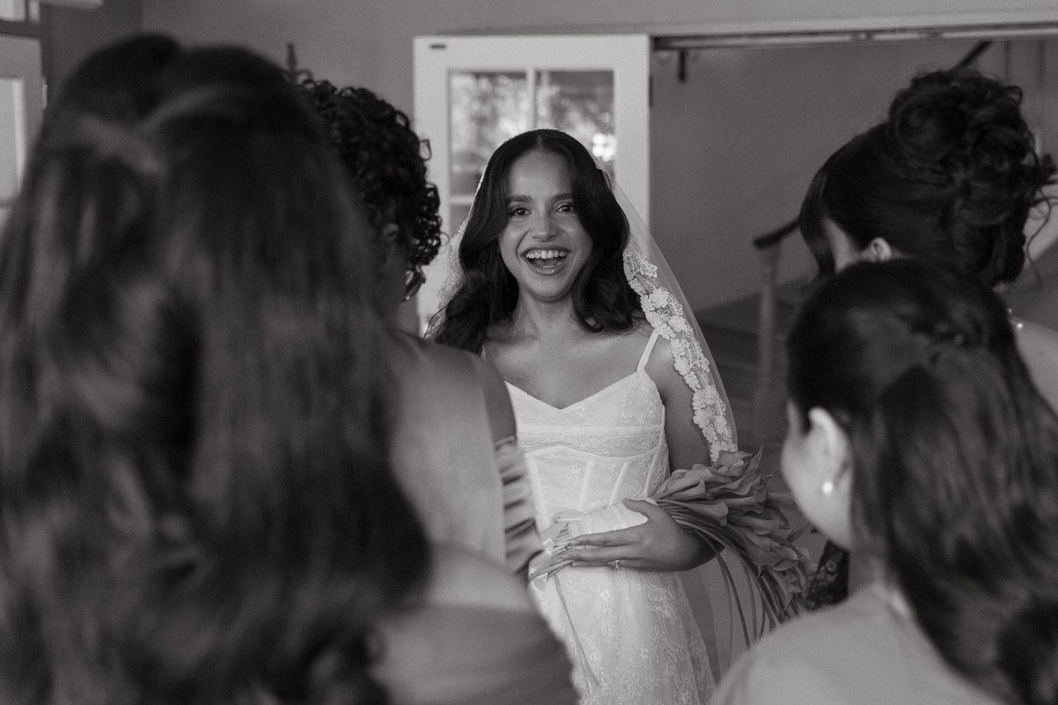 Black and white photo of a smiling bride in a wedding dress and veil surrounded by bridesmaids.