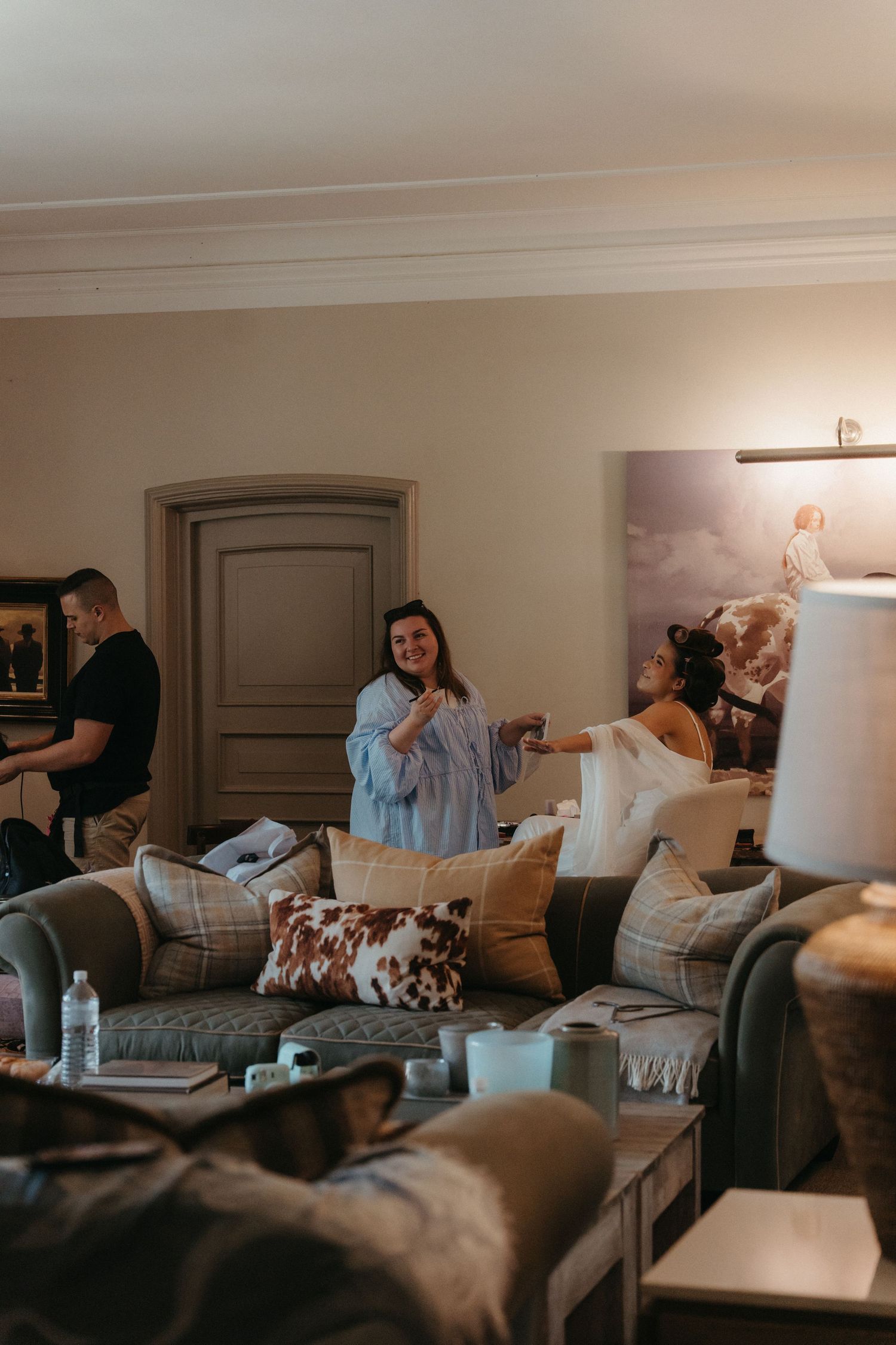 Bride and makeup artist laughing and relaxing in an elegantly decorated living room with plush sofas and warm lighting.
