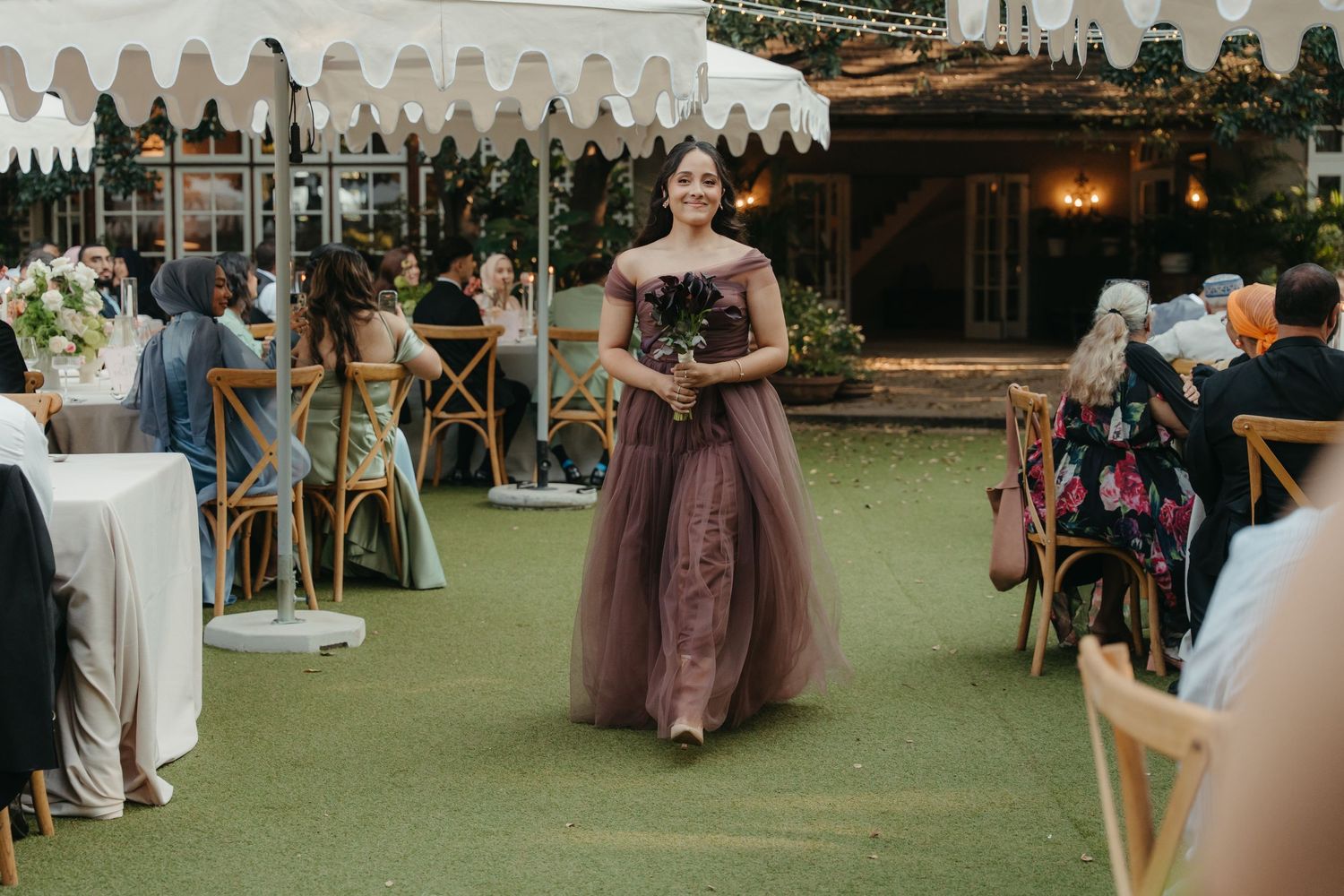 Bridesmaid in purple tulle gown walks down outdoor wedding aisle at elegant garden ceremony.
