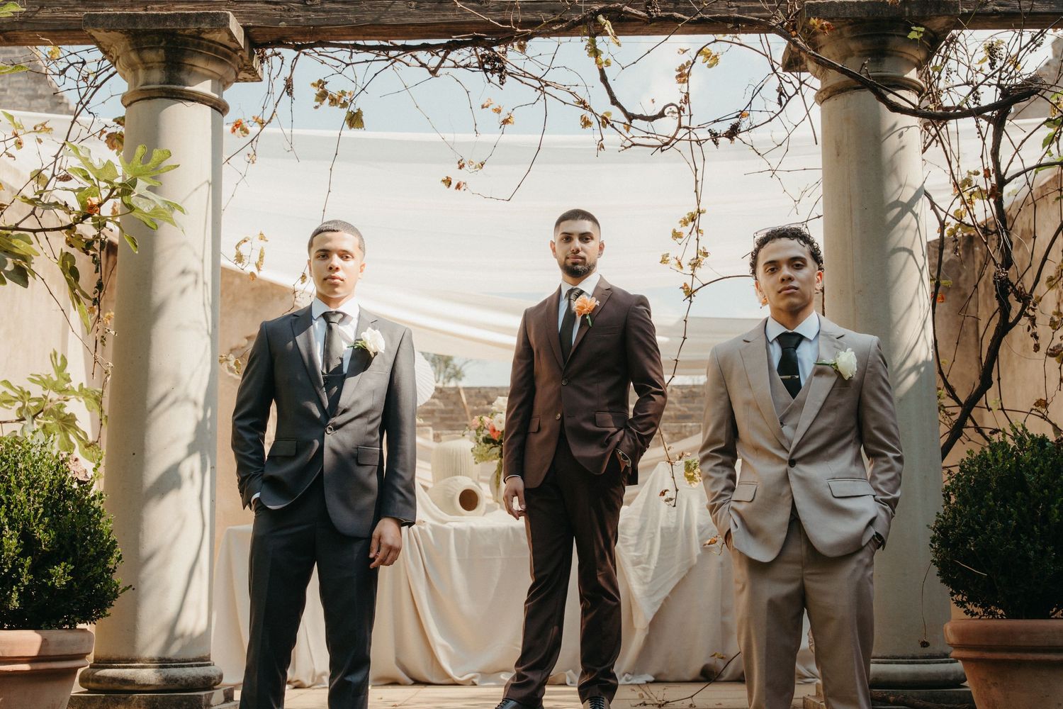 Three groomsmen in suits stand under stone columns at an elegant outdoor wedding venue.