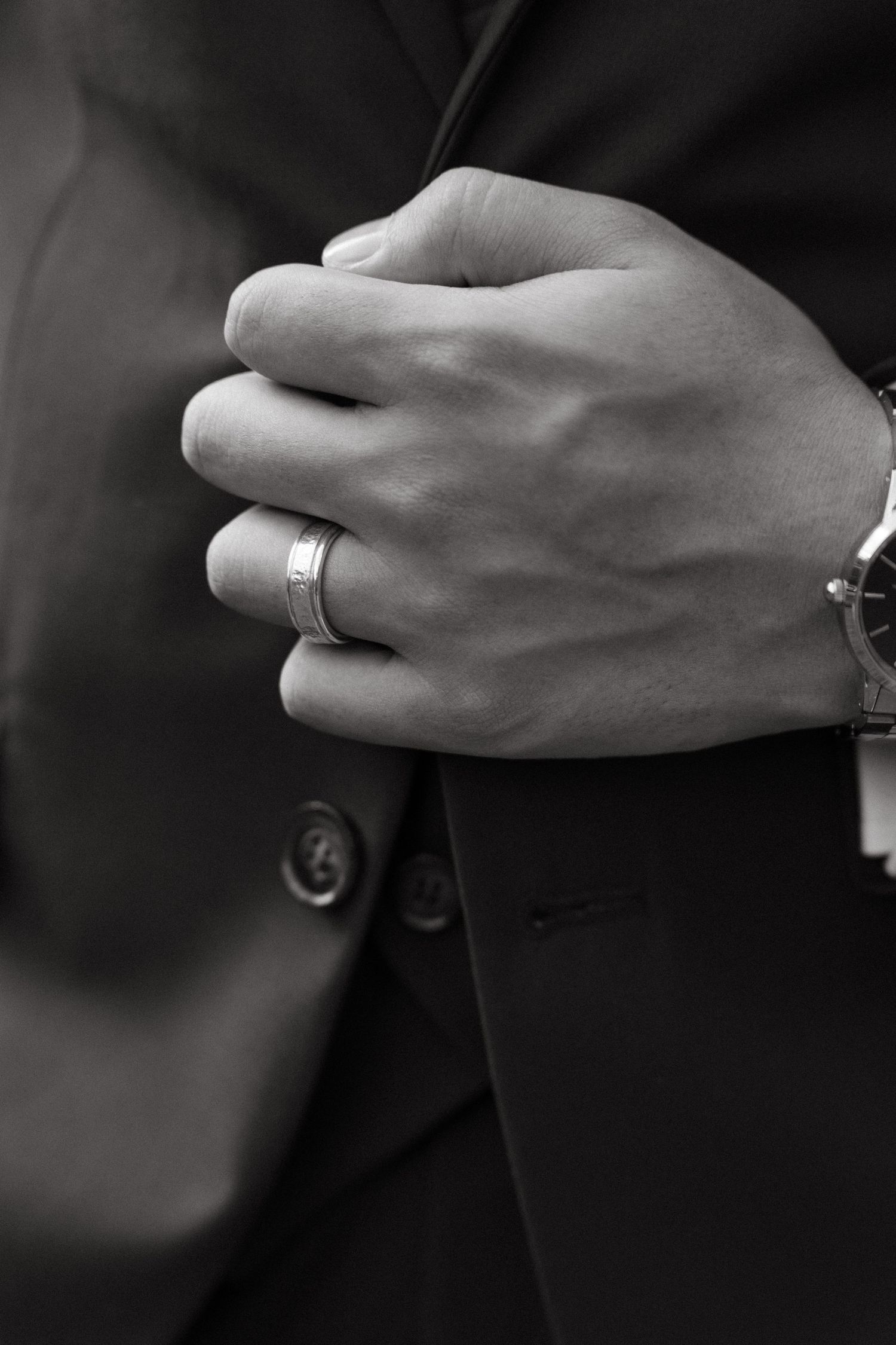 Black and white close-up of a man in a suit with a wedding ring and watch, hands clasped together.