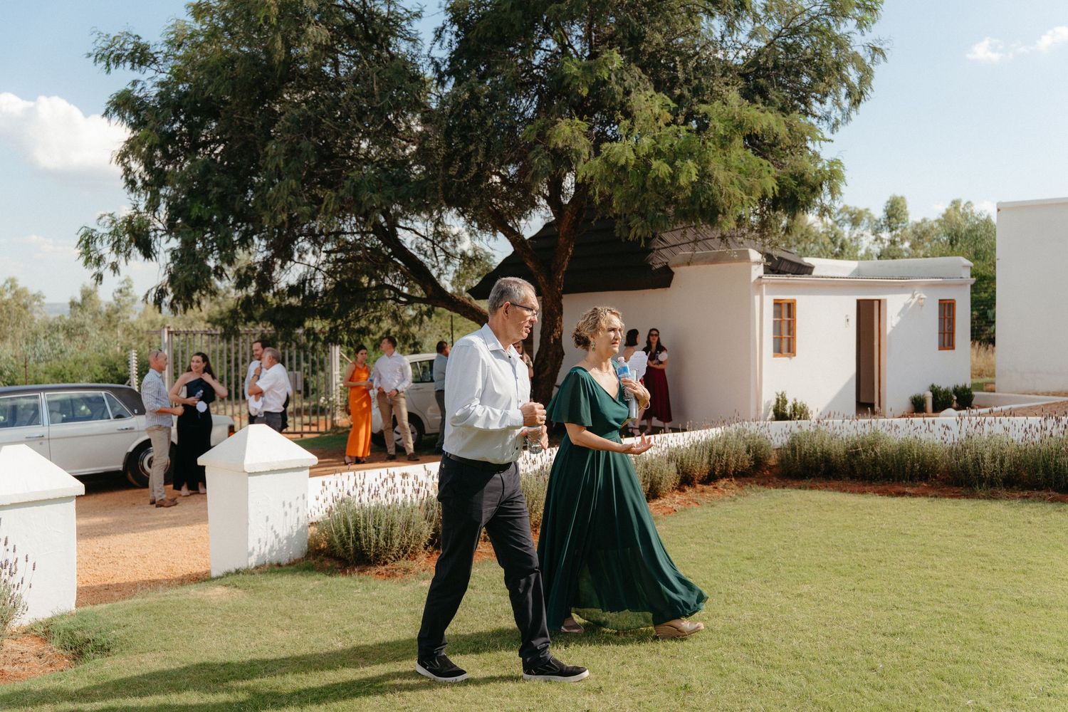 Two people walk arm in arm across a lush green lawn outside a white building on a sunny day.