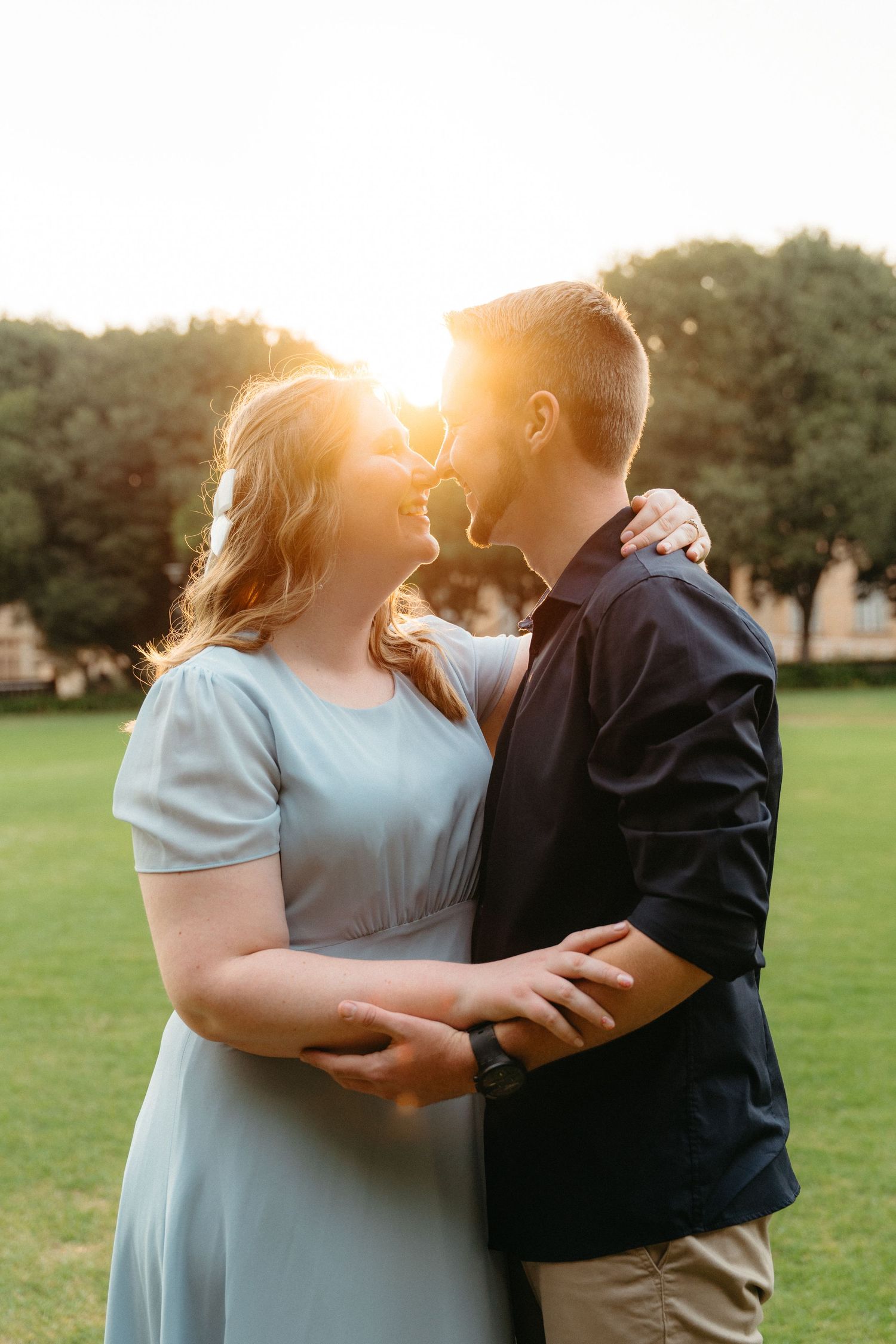 Romantic couple embraces in a grassy field during golden hour sunset with sunlight glowing between them.