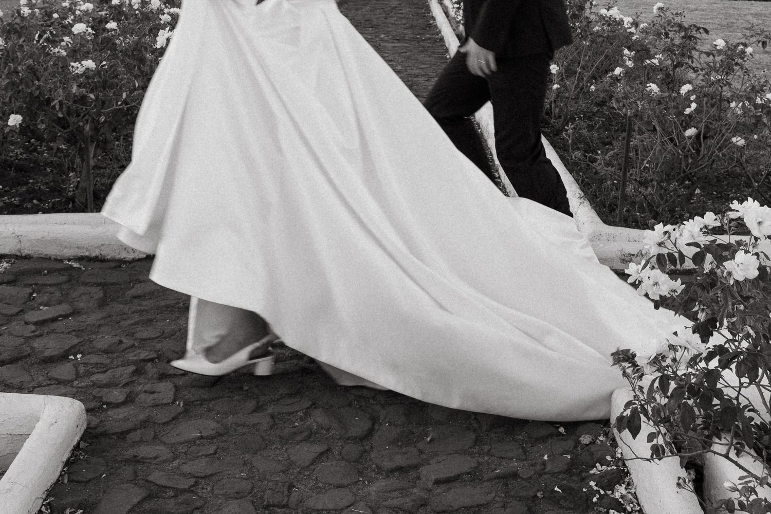 A couple in wedding attire embraces romantically in a garden setting with trees and greenery in the background.