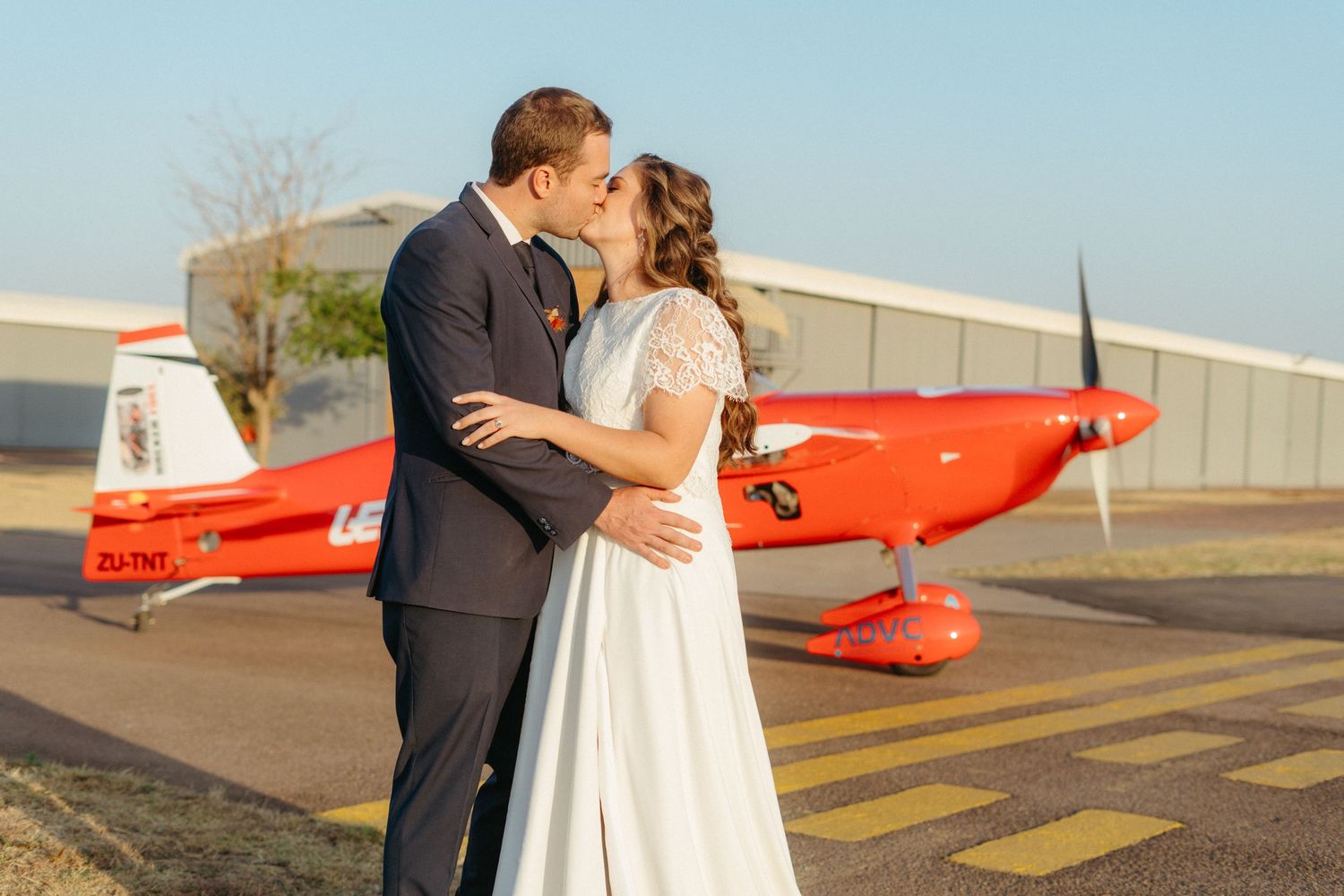 A romantic kiss shared in front of a bright red vintage aircraft on an airfield.