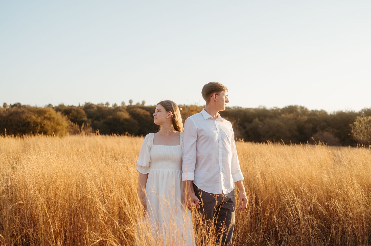 A couple in white clothing walking through tall golden grass during sunset.