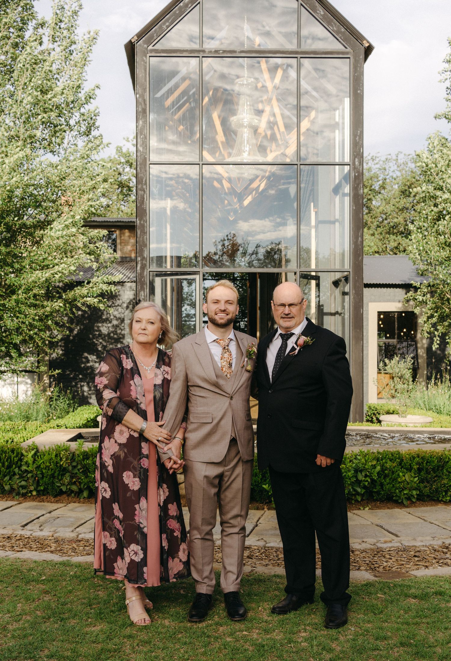 Three people stand together in formal attire in front of a glass greenhouse structure in a garden setting.