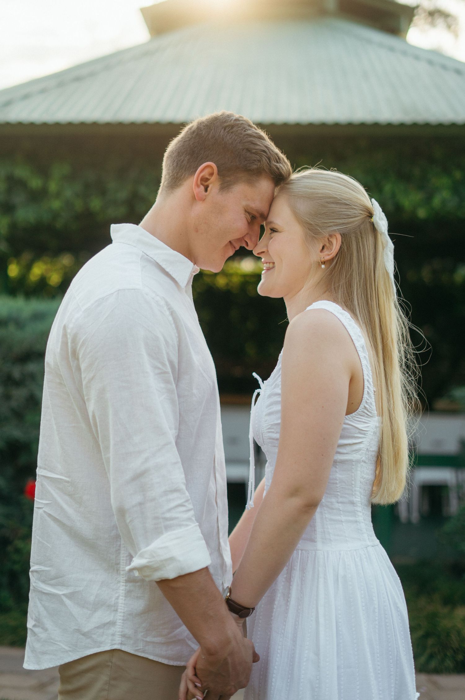 A romantic couple shares an intimate moment together outdoors on a sunny day wearing coordinated white outfits.