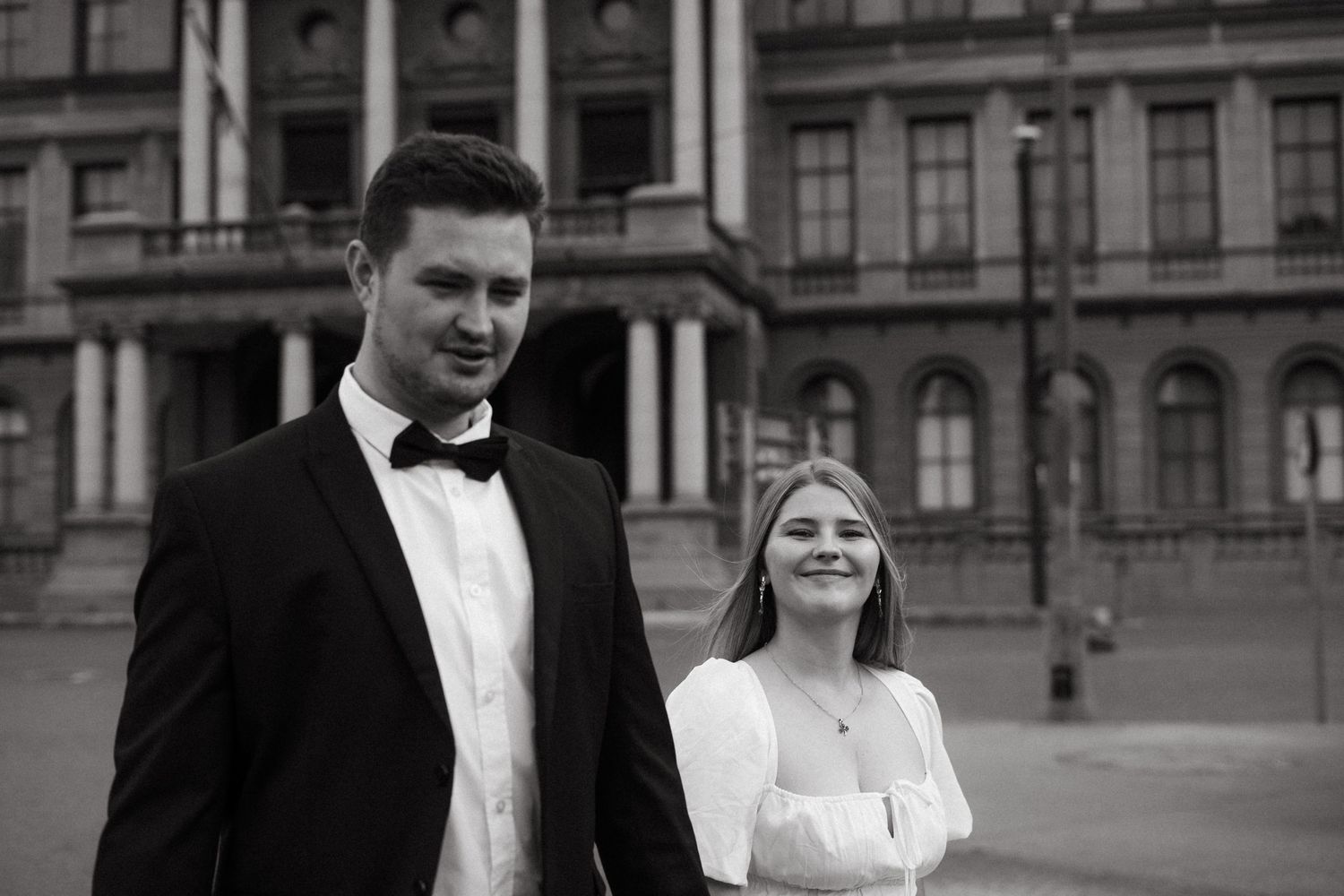 Black and white photo of a celebratory couple in formal attire outside a grand historic building.