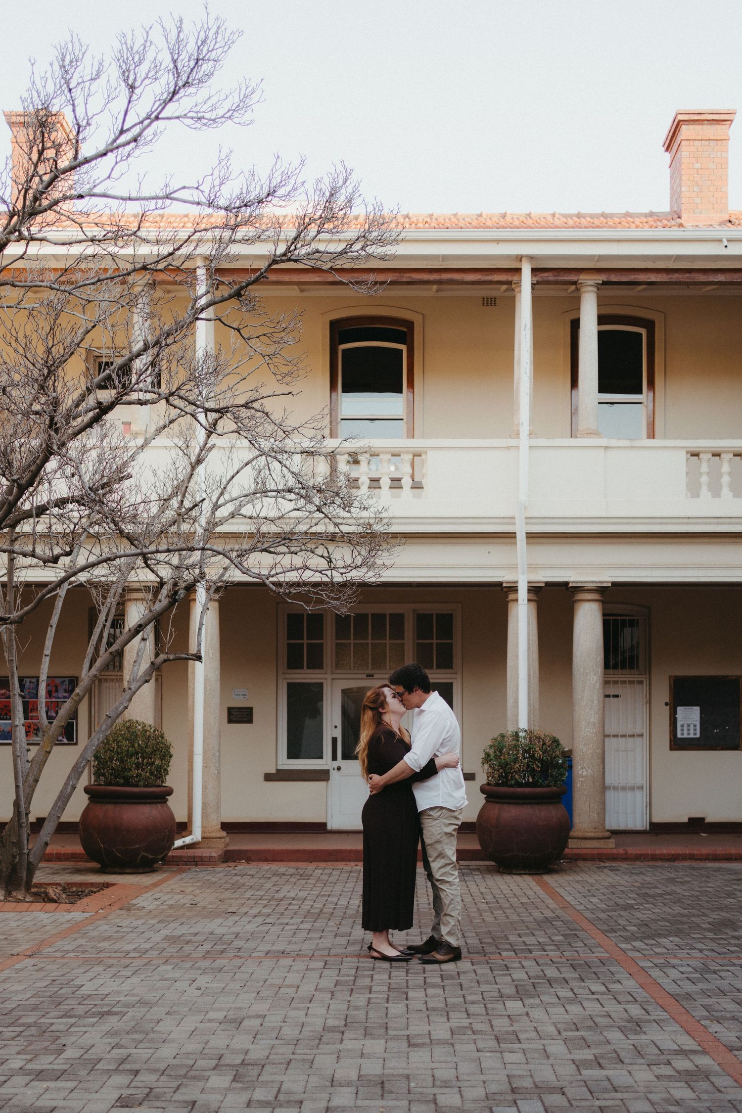 Couple sharing a romantic kiss in front of a charming two-story University of Pretoria heritage building.