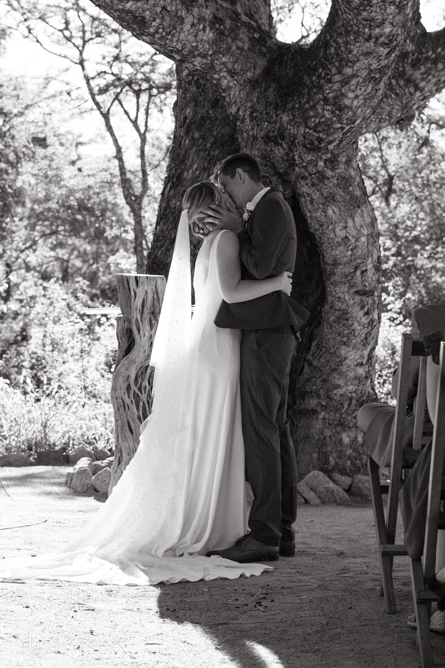 A romantic black and white wedding portrait of a couple embracing under a large tree.