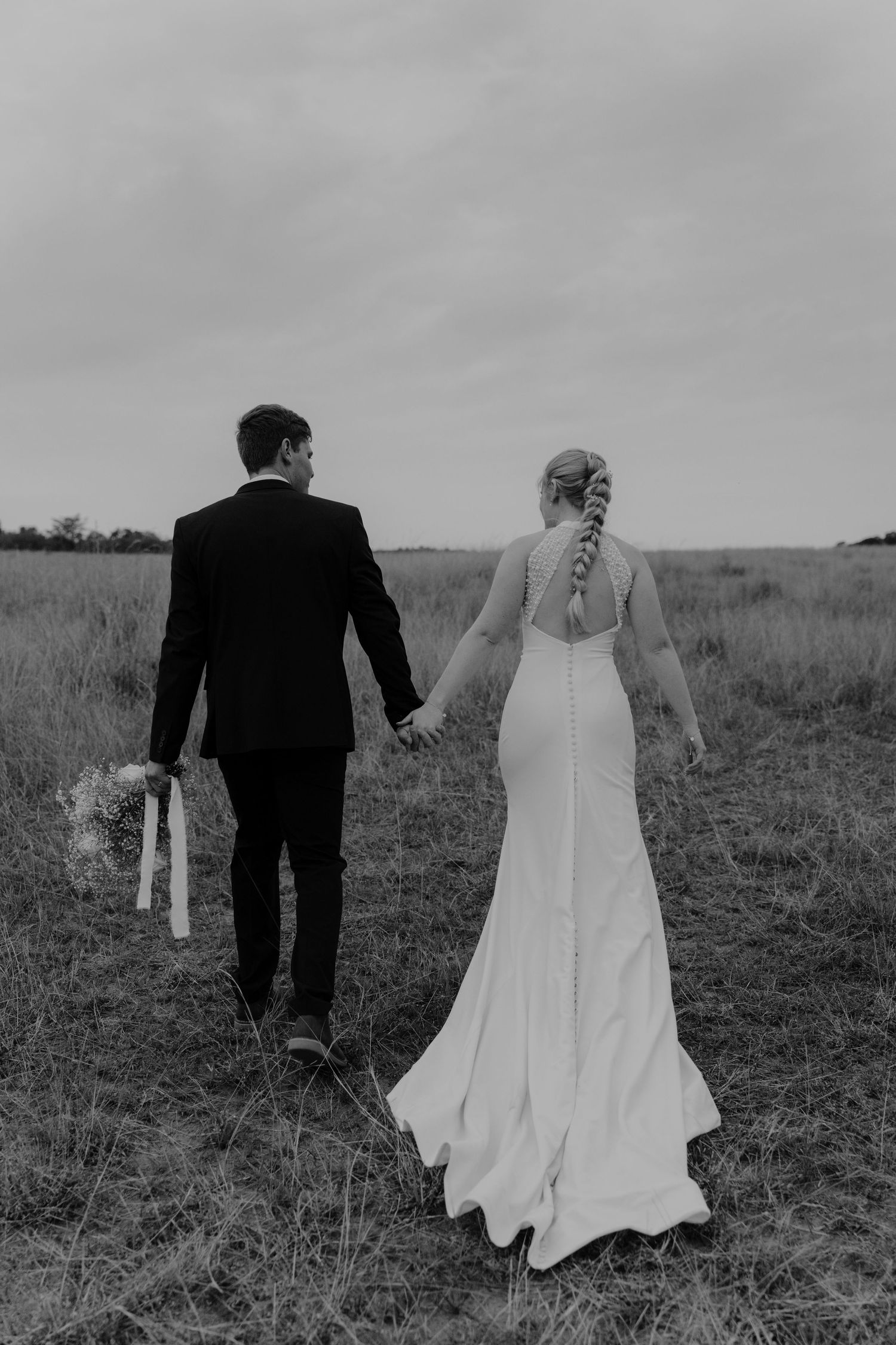 A bride and groom walk through a grassy field holding hands in black and white.