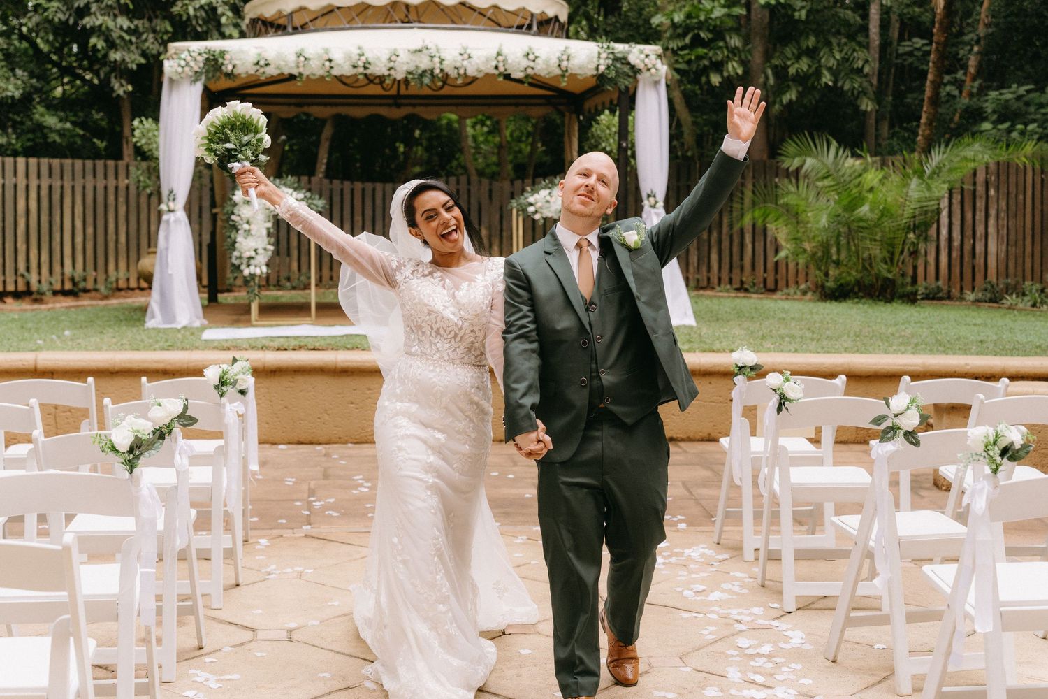 A wedding celebration in an outdoor garden venue with white chairs and decorative floral arrangements.