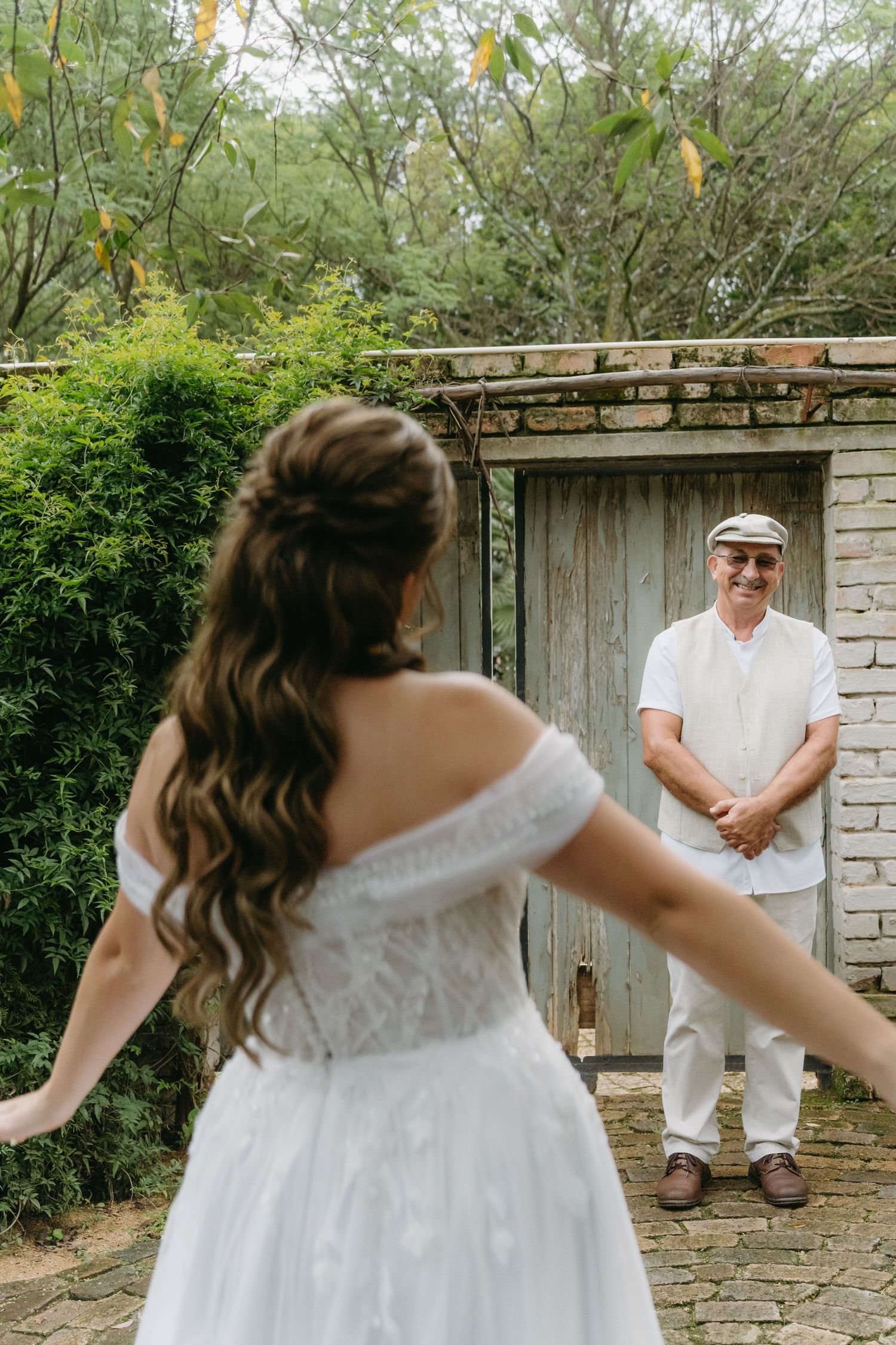 Two people in white outfits share a special first look moment by a rustic wooden door in a garden setting.