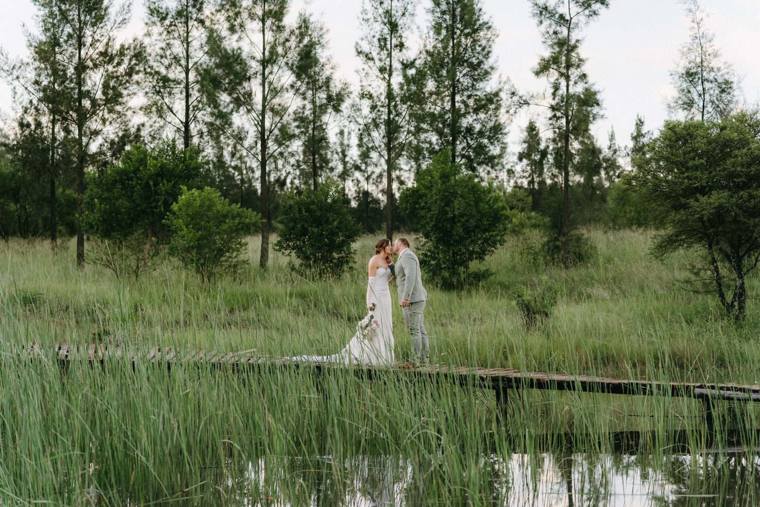 A couple in wedding attire stands on a wooden boardwalk over a serene pond surrounded by tall grass.