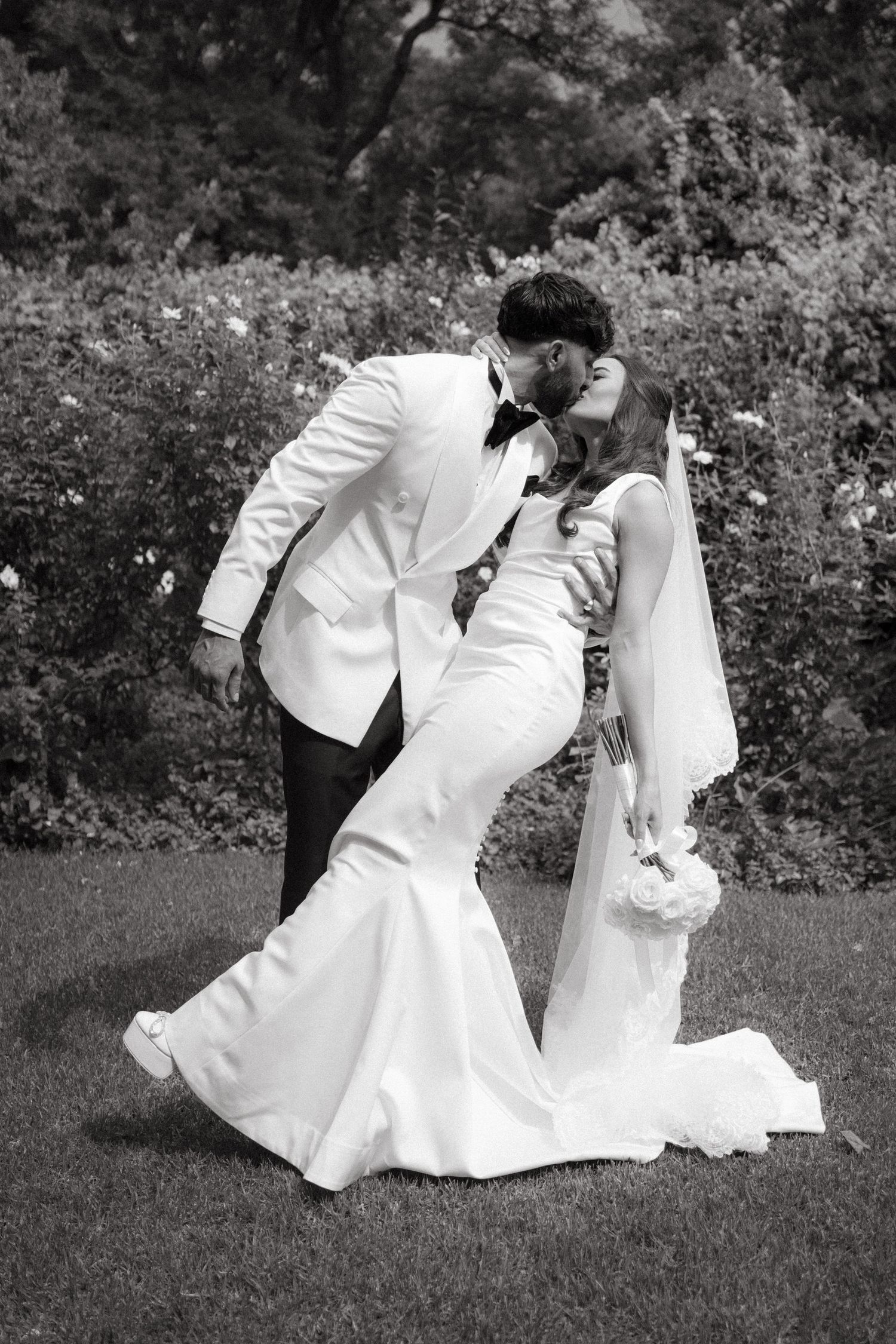 A romantic black and white wedding photo showing a couple in white attire sharing an intimate dip pose on a lawn.