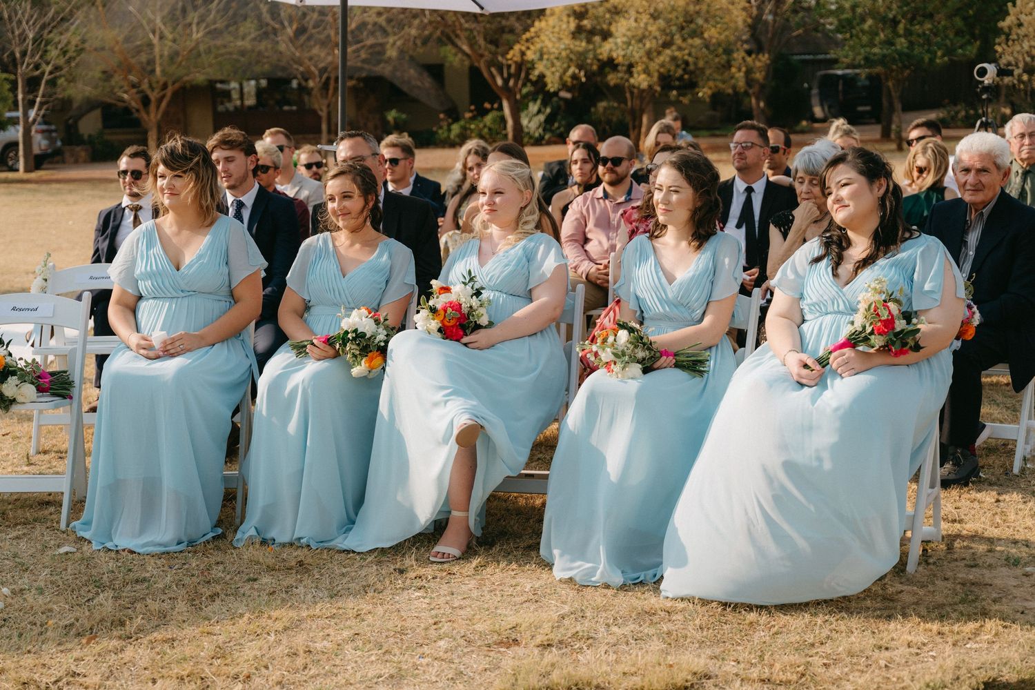 Bridesmaids in light blue dresses seated together at an outdoor wedding ceremony.