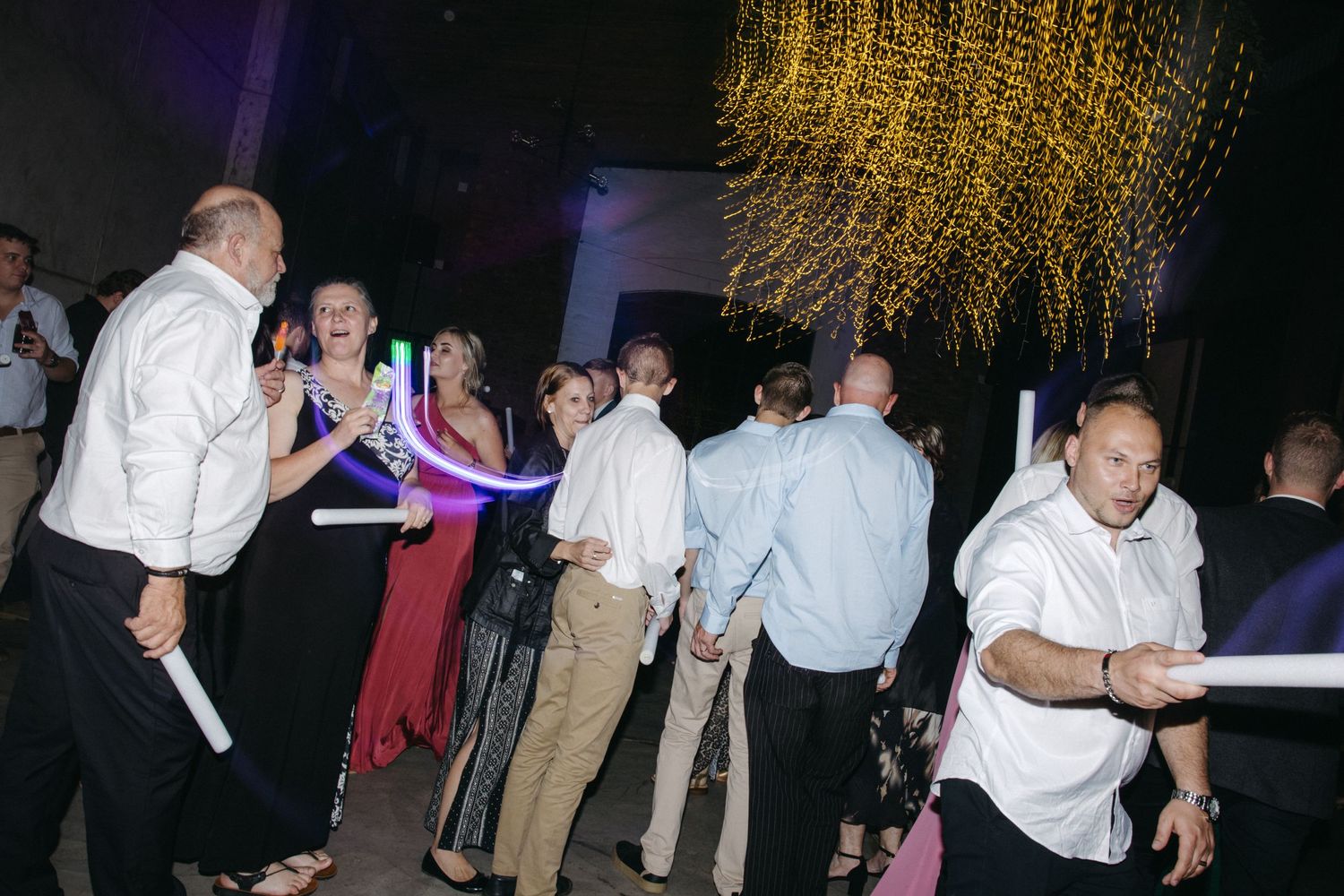Wedding reception guests dancing with glow sticks under decorative hanging lights.