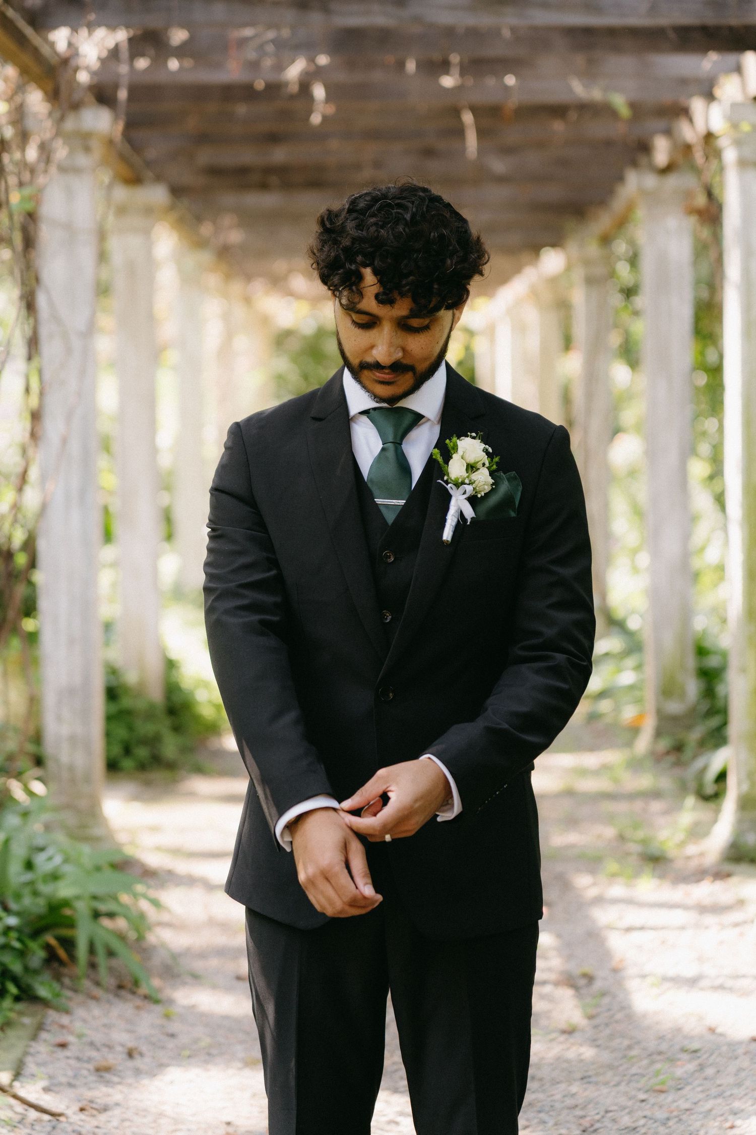 Person in a black suit adjusts their boutonniere during an outdoor wedding photoshoot under wooden beams.