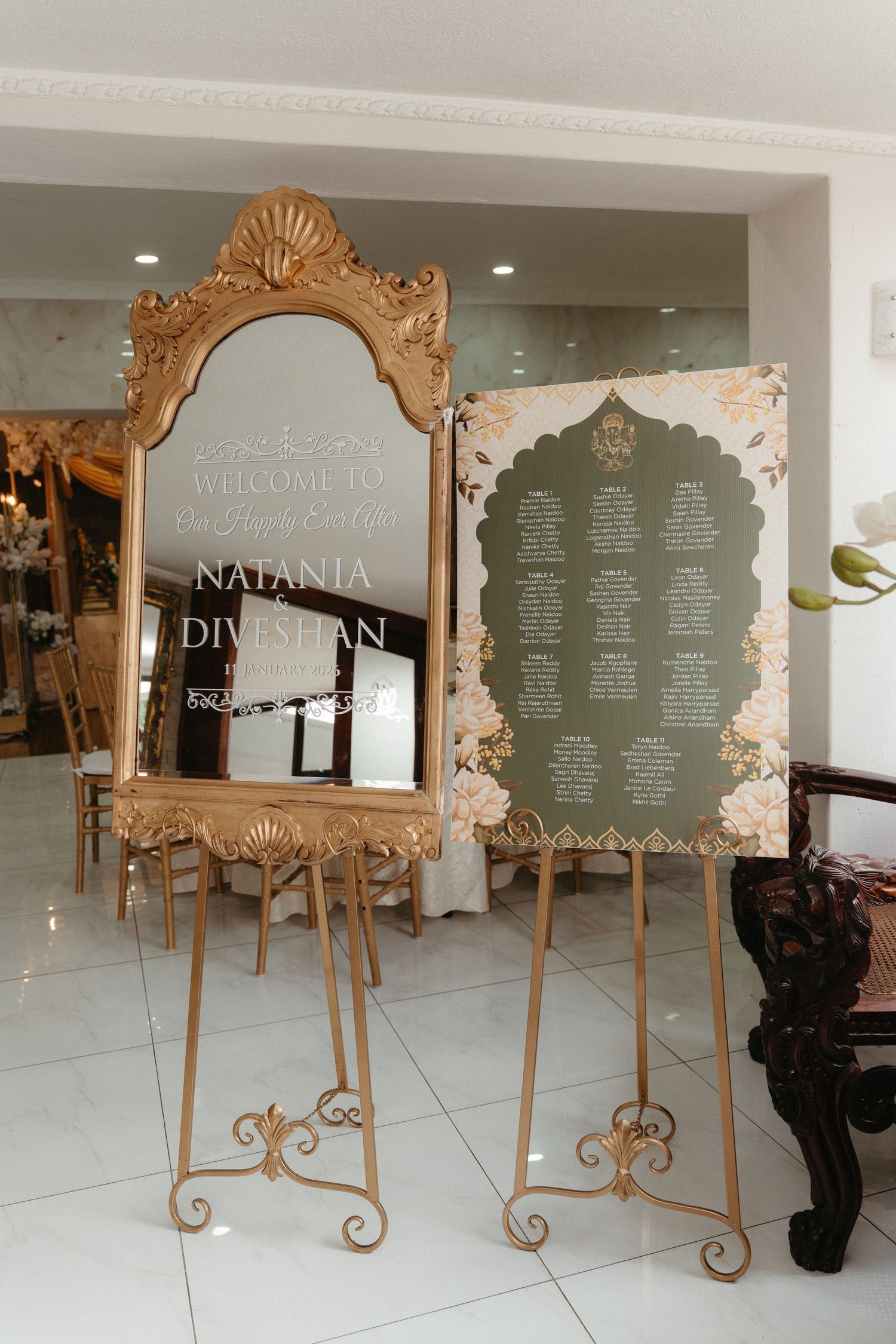 Ornate gold-framed mirrors with seating charts displayed on sage green backgrounds stand on elegant easels at a wedding venue.