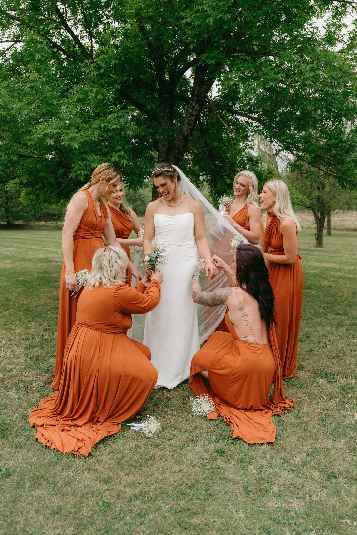 Bridesmaids in orange dresses sitting on grass with bride in white gown.