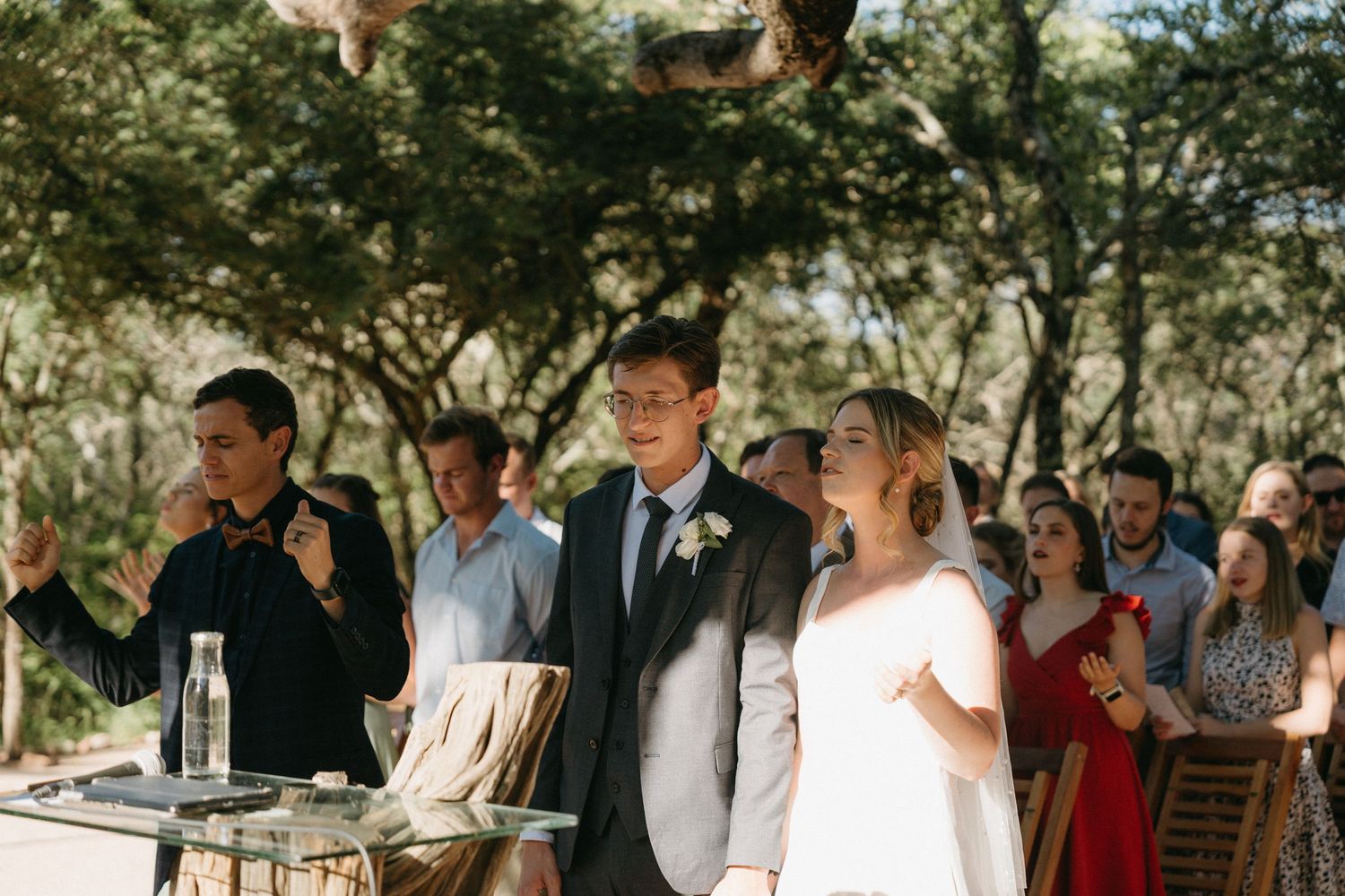 An outdoor wedding ceremony in progress with guests seated in a natural wooded setting.