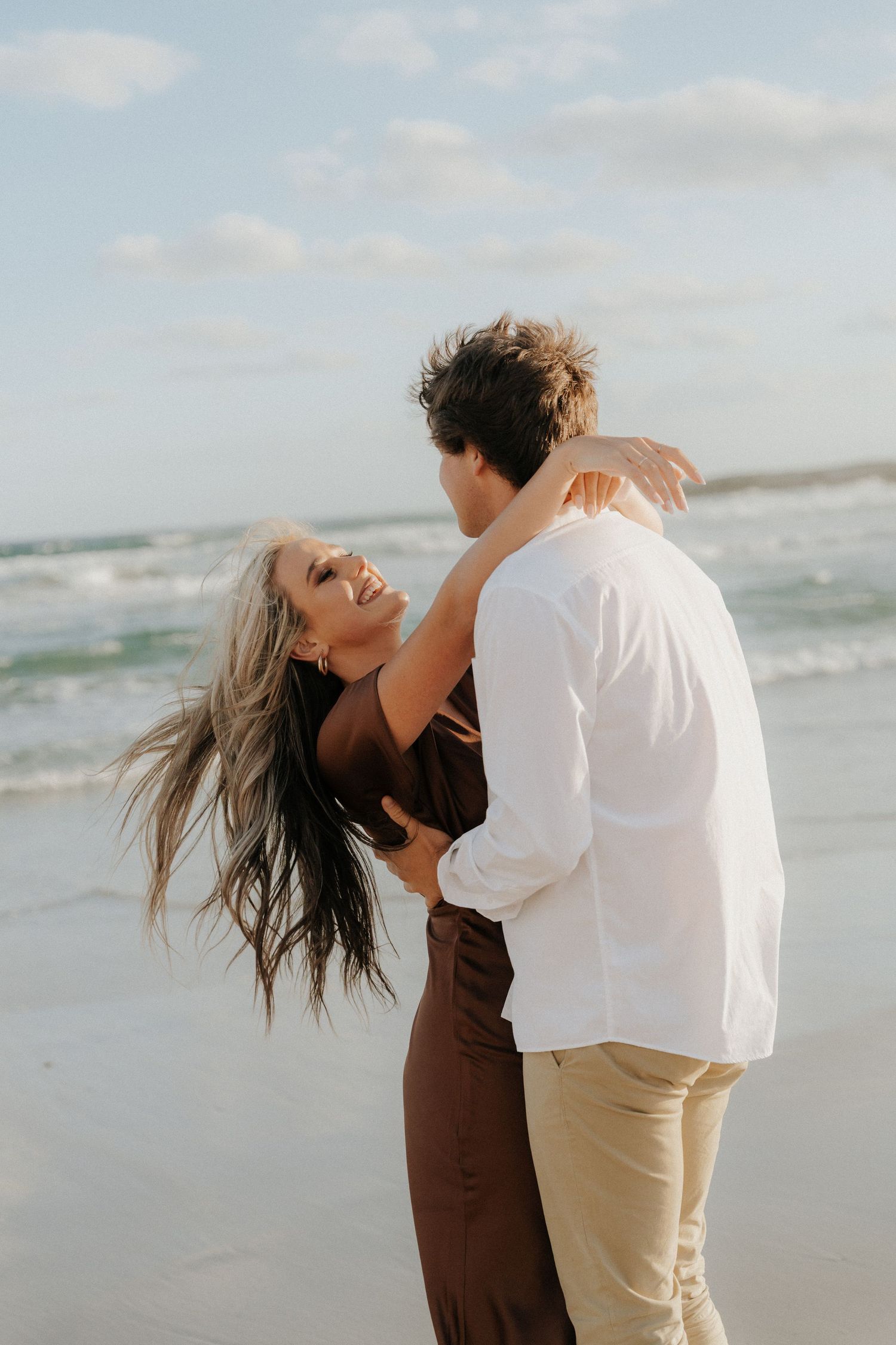 A couple embraces and laughs together on a breezy beach, woman in brown dress with flowing hair, man in white shirt.