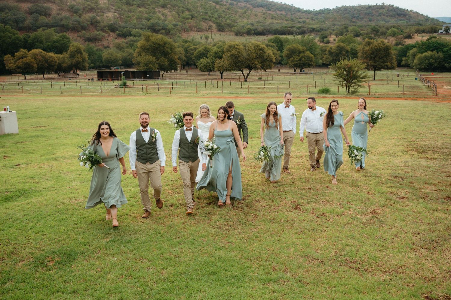 Wedding party in light blue and neutral attire walks joyfully across a grassy field with mountains in the background.