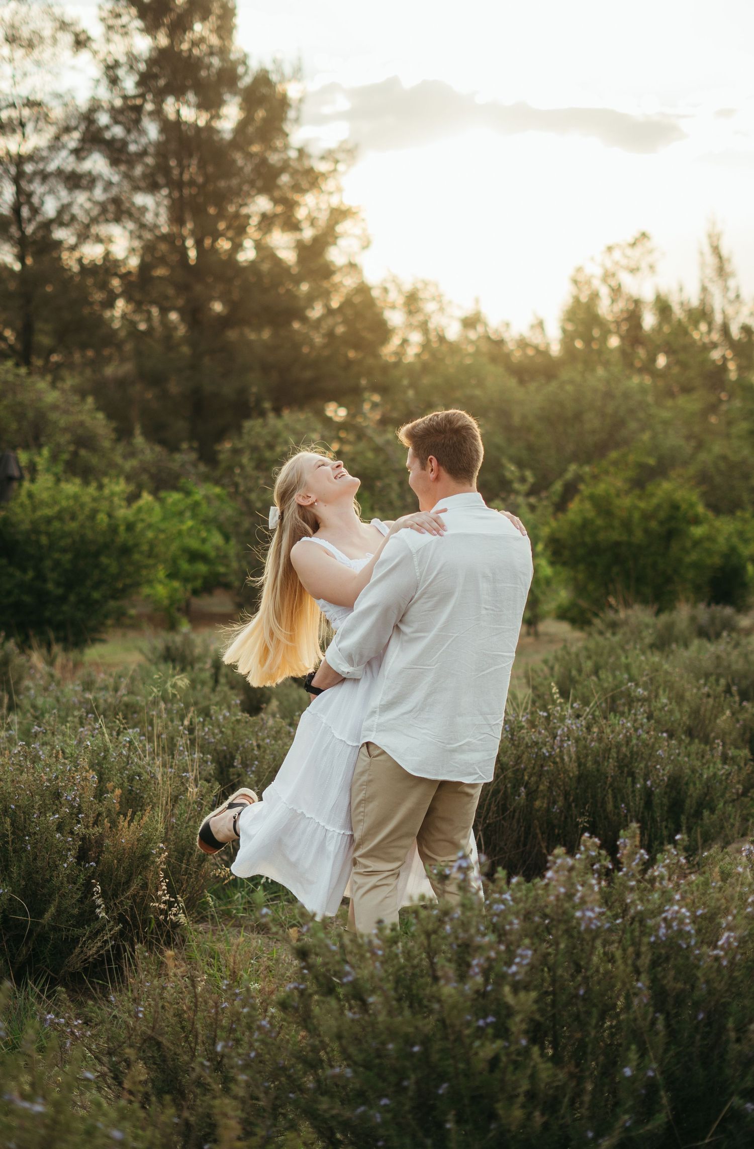 A couple dances together in a wildflower field at sunset with trees silhouetted in the background.
