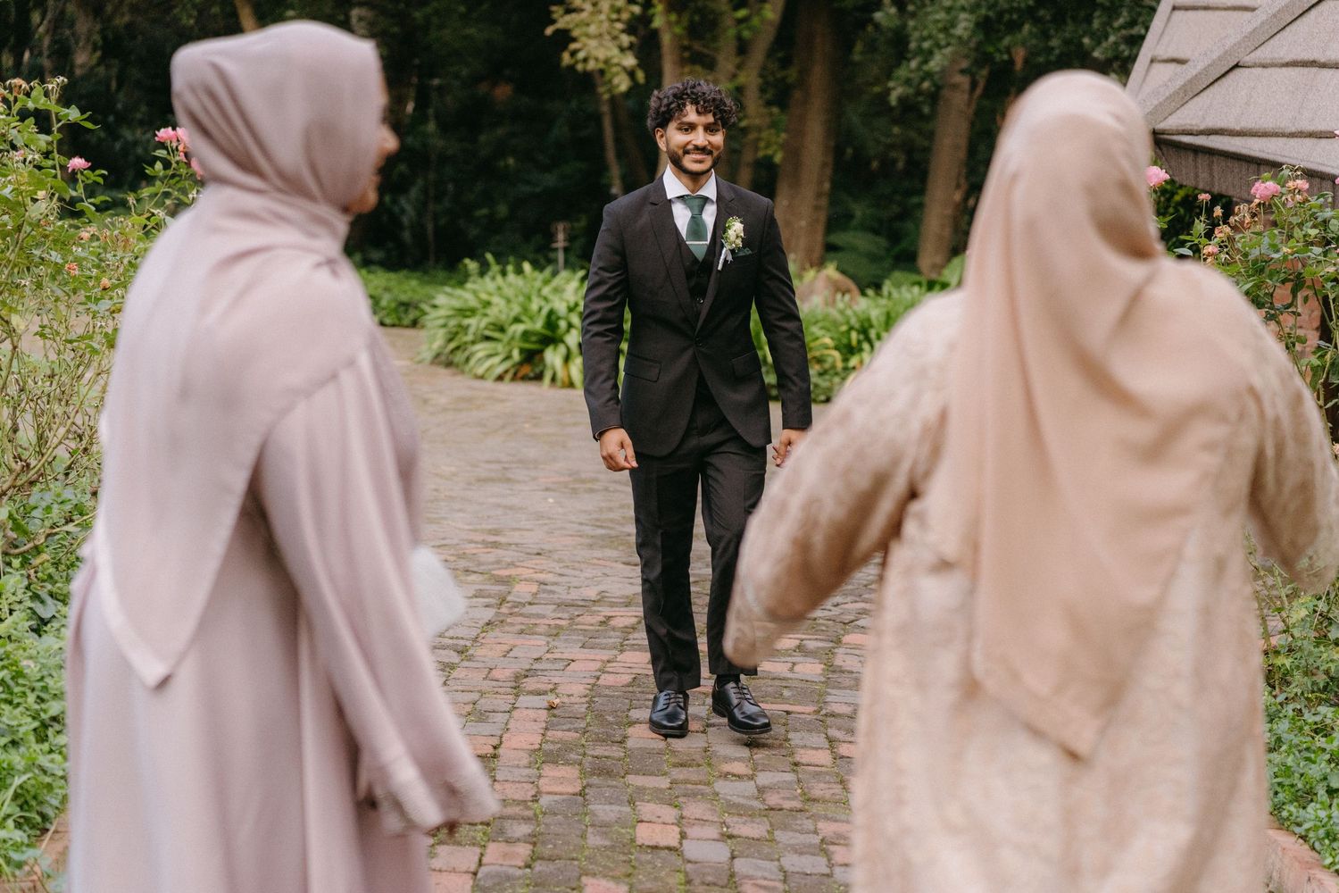 Wedding attendees in traditional hijabs greeting the groom on a garden pathway.