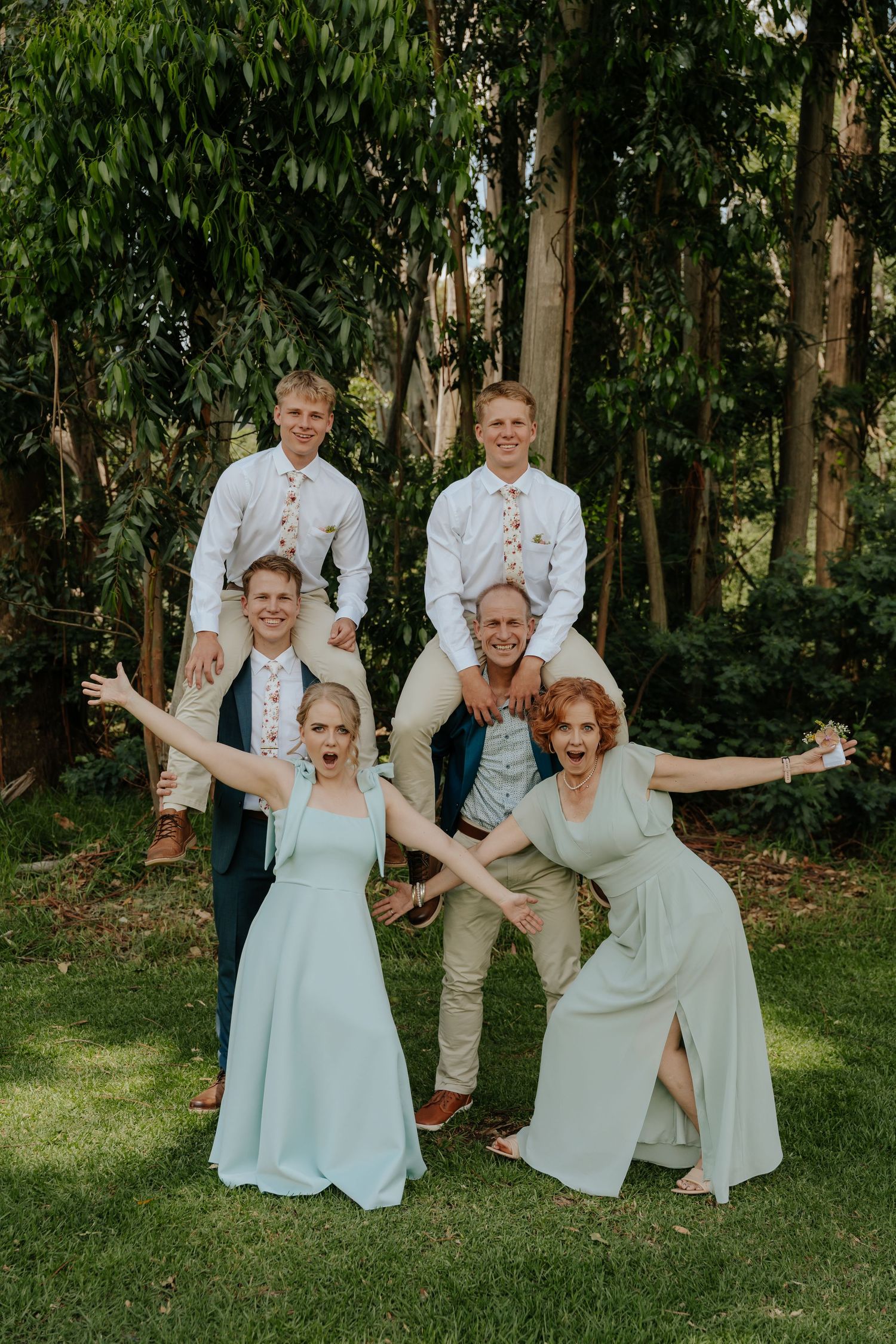 A wedding party in white and light blue attire poses playfully on a grassy lawn with trees in the background.