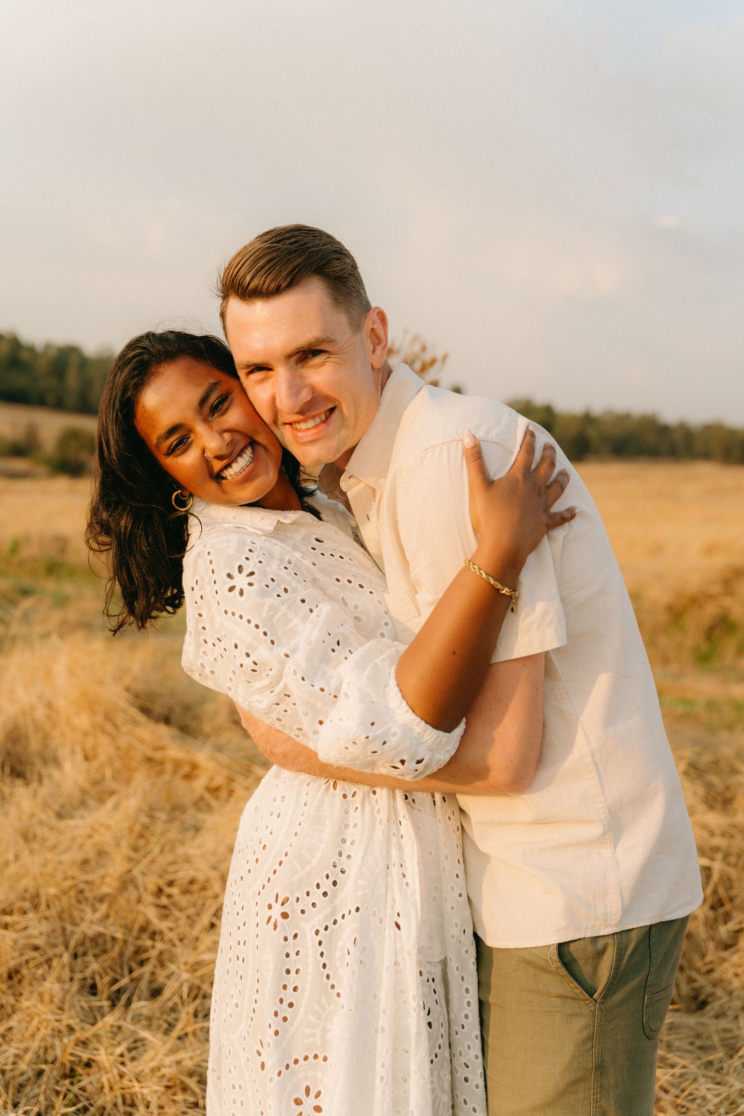A couple embraces lovingly in a golden wheat field during sunset, with the woman wearing a white eyelet dress.