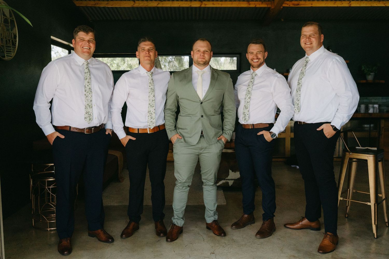 Five groomsmen in formal attire pose together in a rustic indoor venue for a wedding photo.