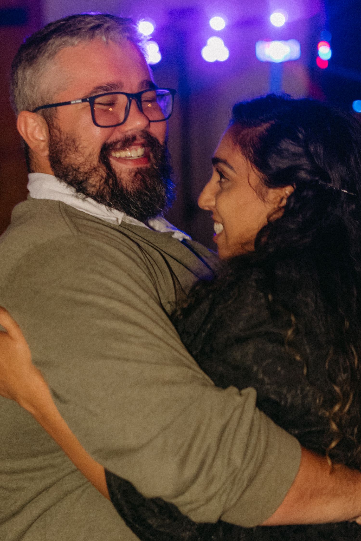 A couple embraces and smiles during a nighttime event with purple stage lighting in the background.