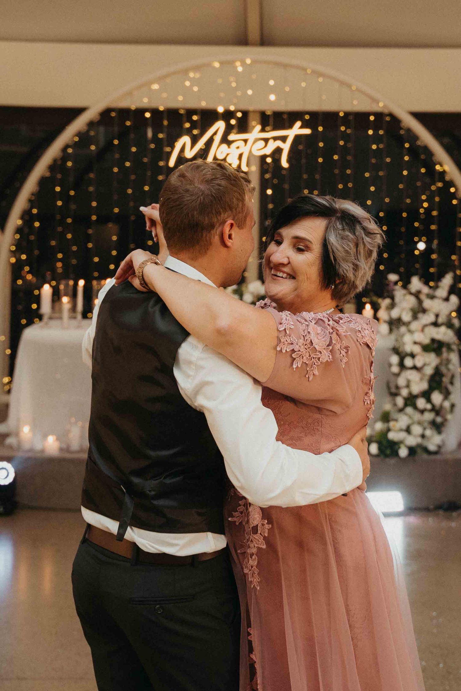 A couple shares a joyful dance at a wedding reception with a glowing Mostert sign and fairy lights backdrop.
