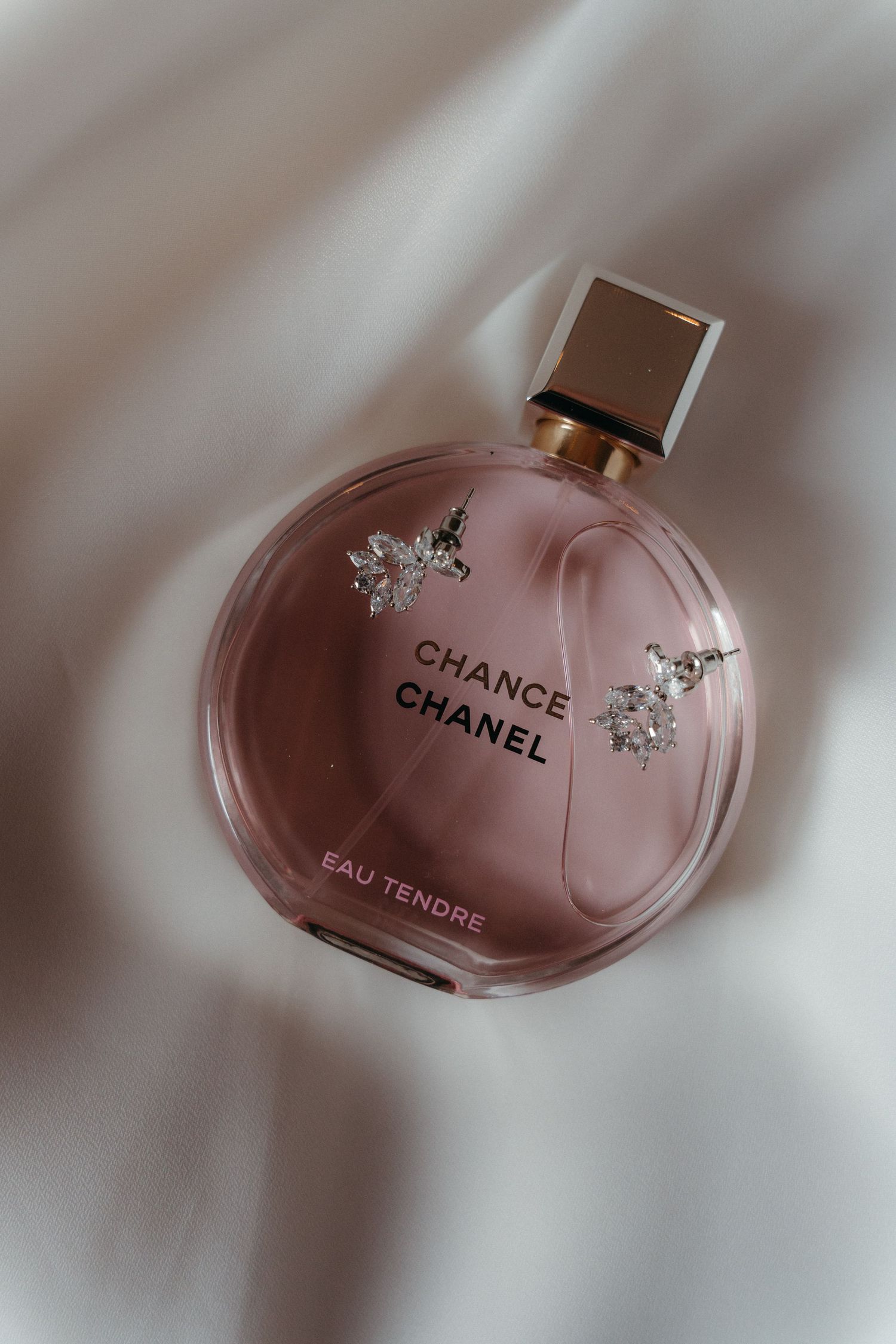 Chanel Chance Eau Tendre pink round perfume bottle with gold cap and crystal flower accents on white satin fabric.