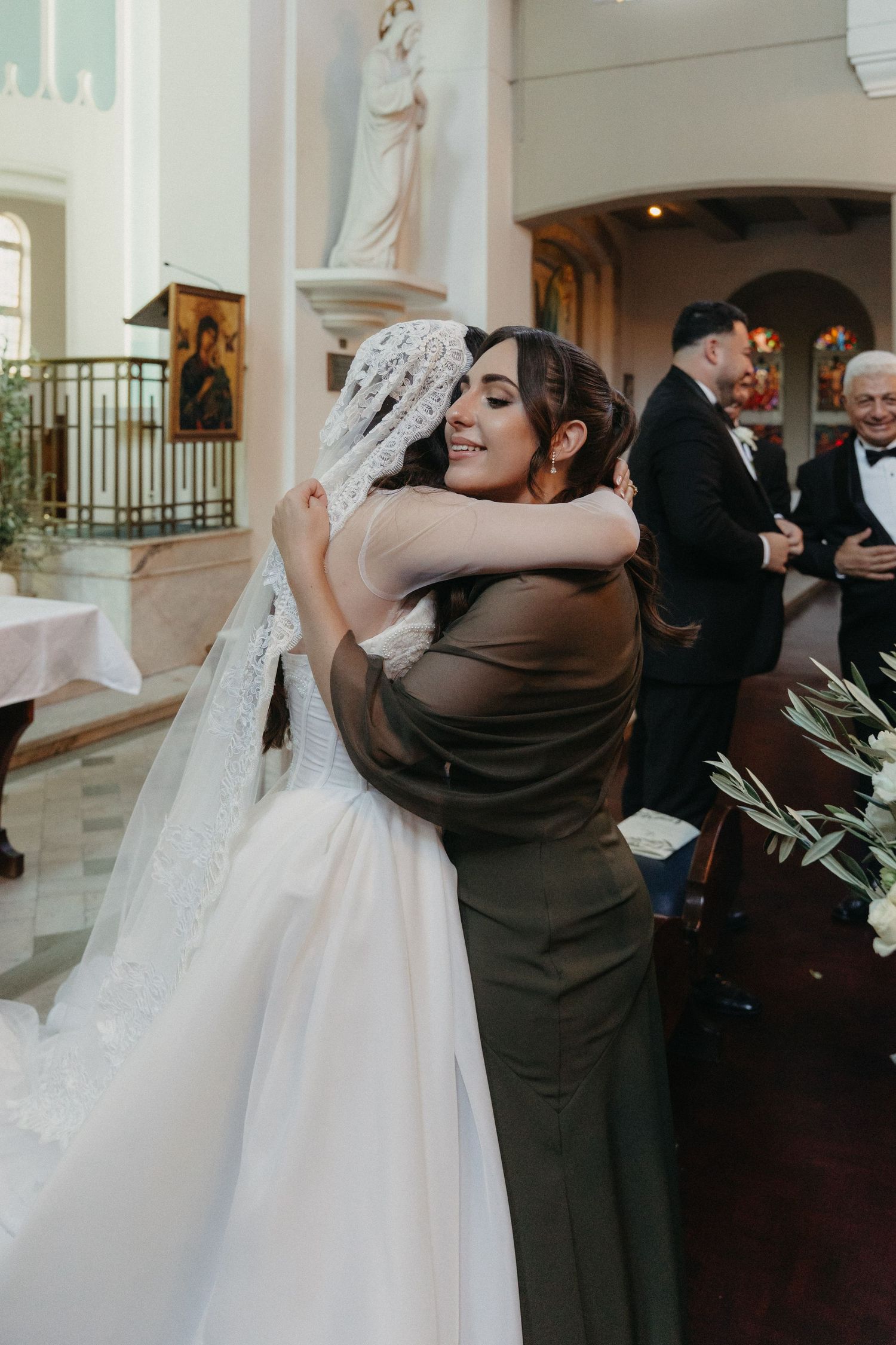 A smiling bride in a lace veil embraces a guest in an olive dress during Rosebank Catholic Church wedding ceremony.
