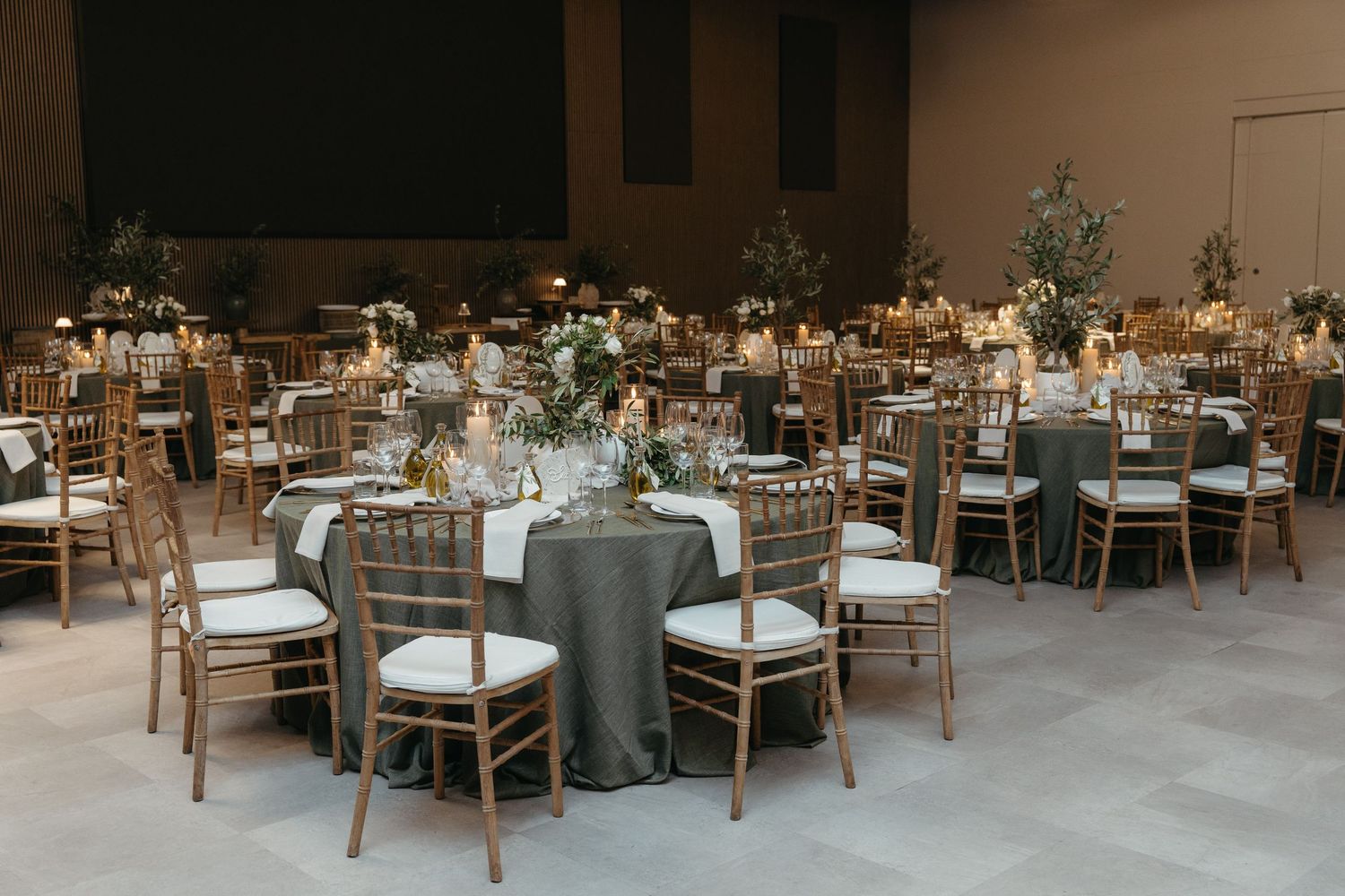 Elegant wedding reception hall with round tables, gold chiavari chairs, sage green linens, and lush floral centerpieces.