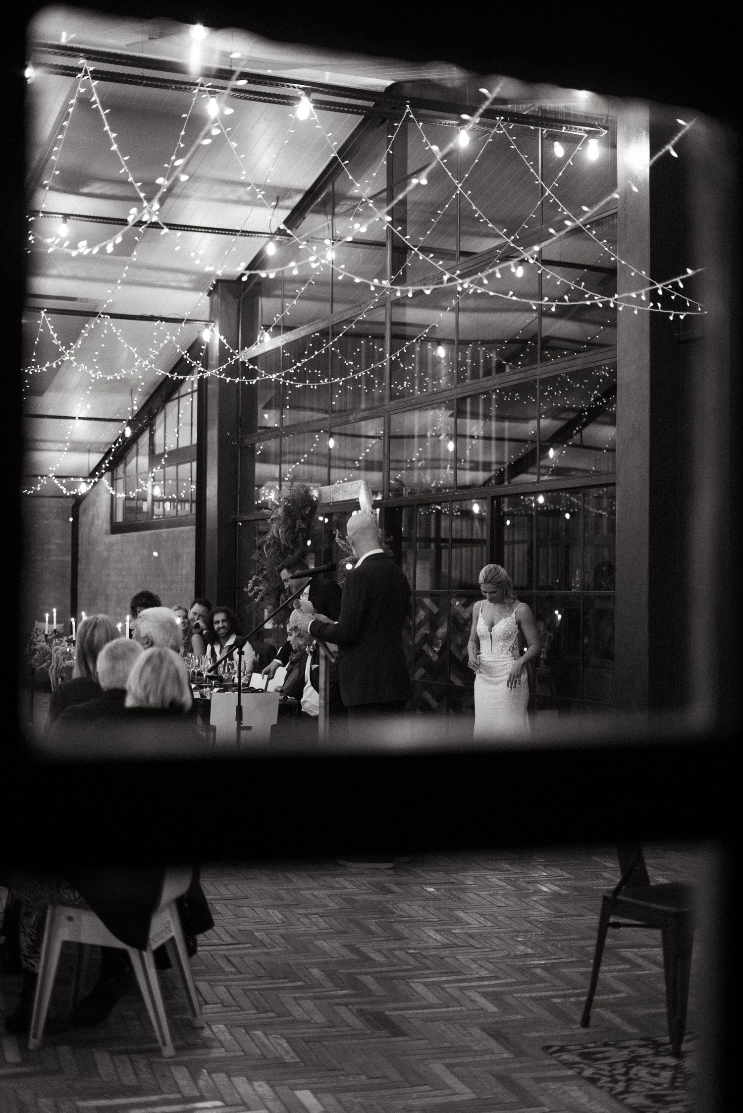 Black and white wedding reception photo through window with string lights and couple standing before seated guests.
