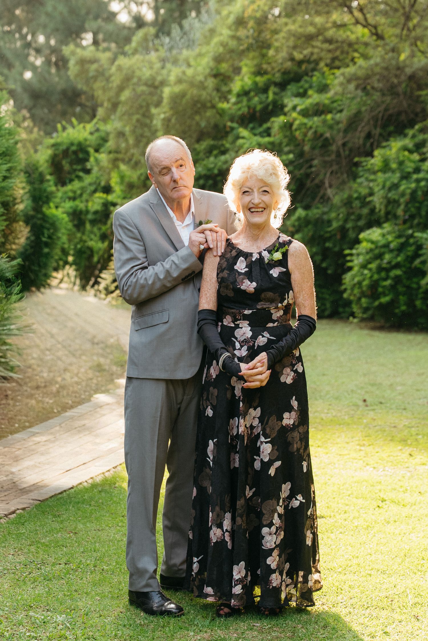 Elderly couple posing outdoors in formal attire, woman in floral gown, man in gray suit, lush green garden background.