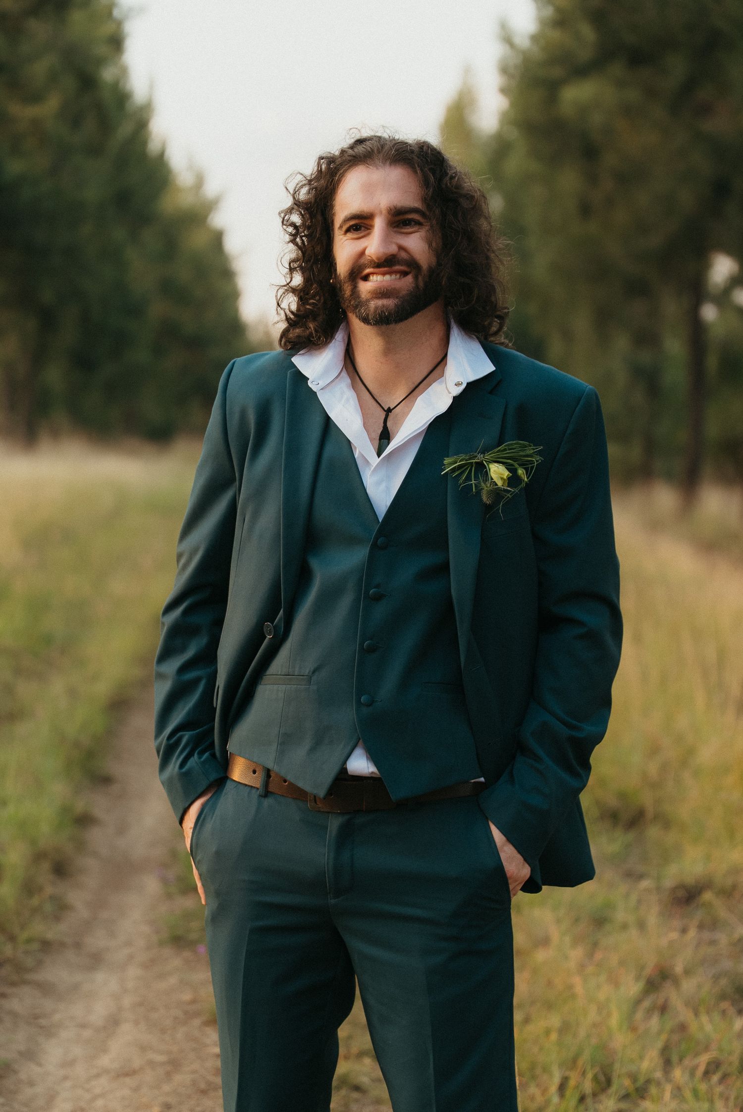 Smiling groom with curly hair in a dark green suit with boutonniere stands on a path surrounded by trees.
