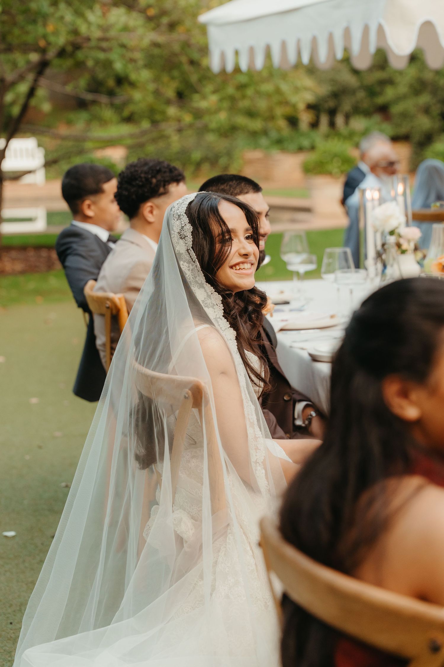 Smiling bride in lace veil seated at outdoor wedding reception table with guests in background.